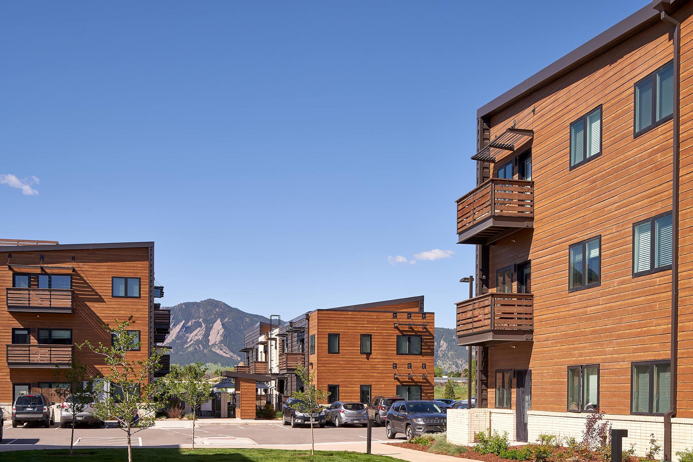 Modern multi-family residential buildings with wood paneling and balconies, situated in a landscaped area with parked cars. The background features a clear blue sky and mountains, creating a scenic environment. The architecture reflects contemporary design elements.