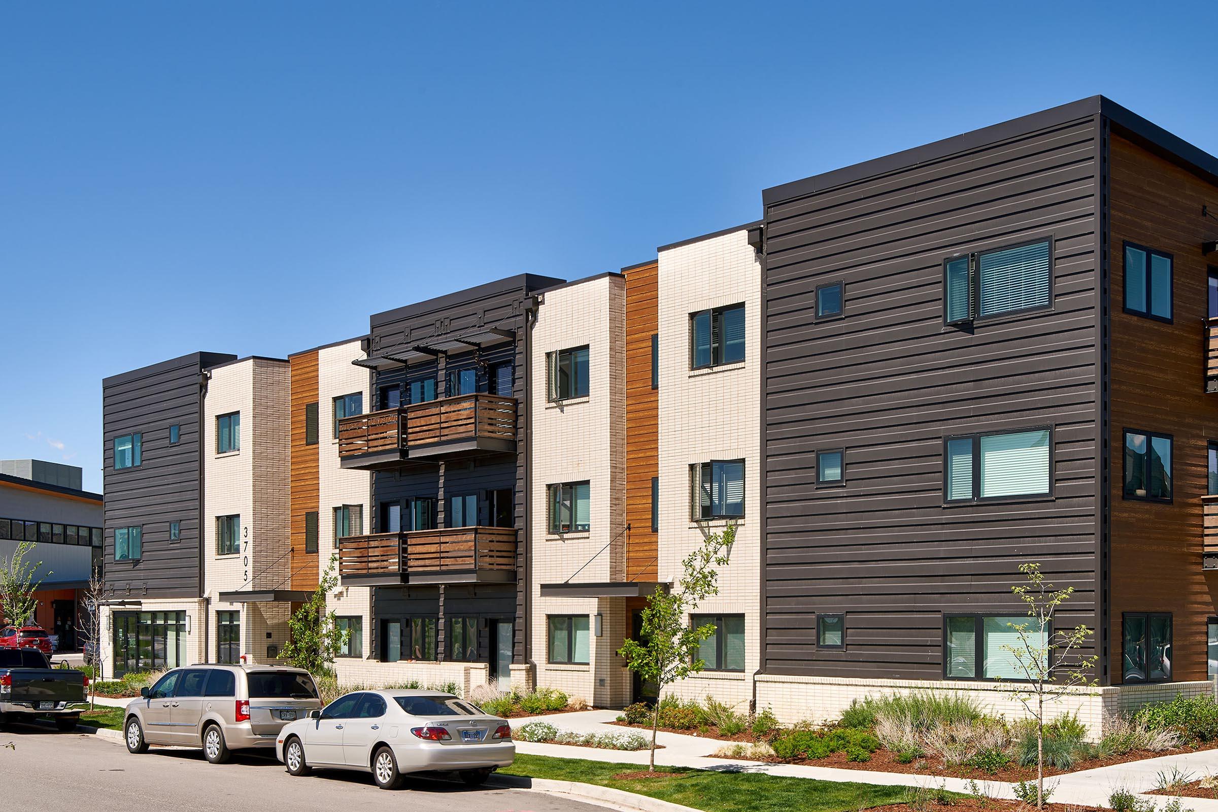 Modern apartment building featuring a mix of materials, including sleek dark panels and light brick. The structure has multiple levels, balconies, and large windows. Surrounding the building are neatly landscaped areas, with parked cars in front and a clear blue sky overhead.