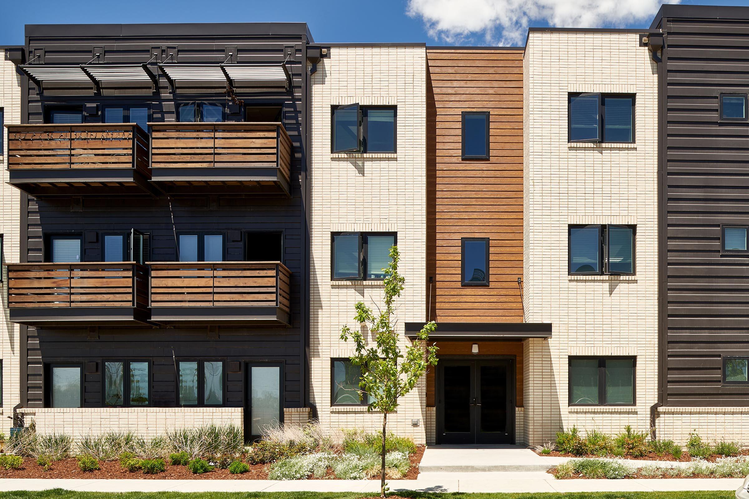 Modern multi-unit building with a mix of dark and light exterior materials. Features include wooden balconies, multiple windows, and a symmetrical design. The front entrance is flanked by small trees and neatly landscaped greenery, creating an inviting atmosphere.