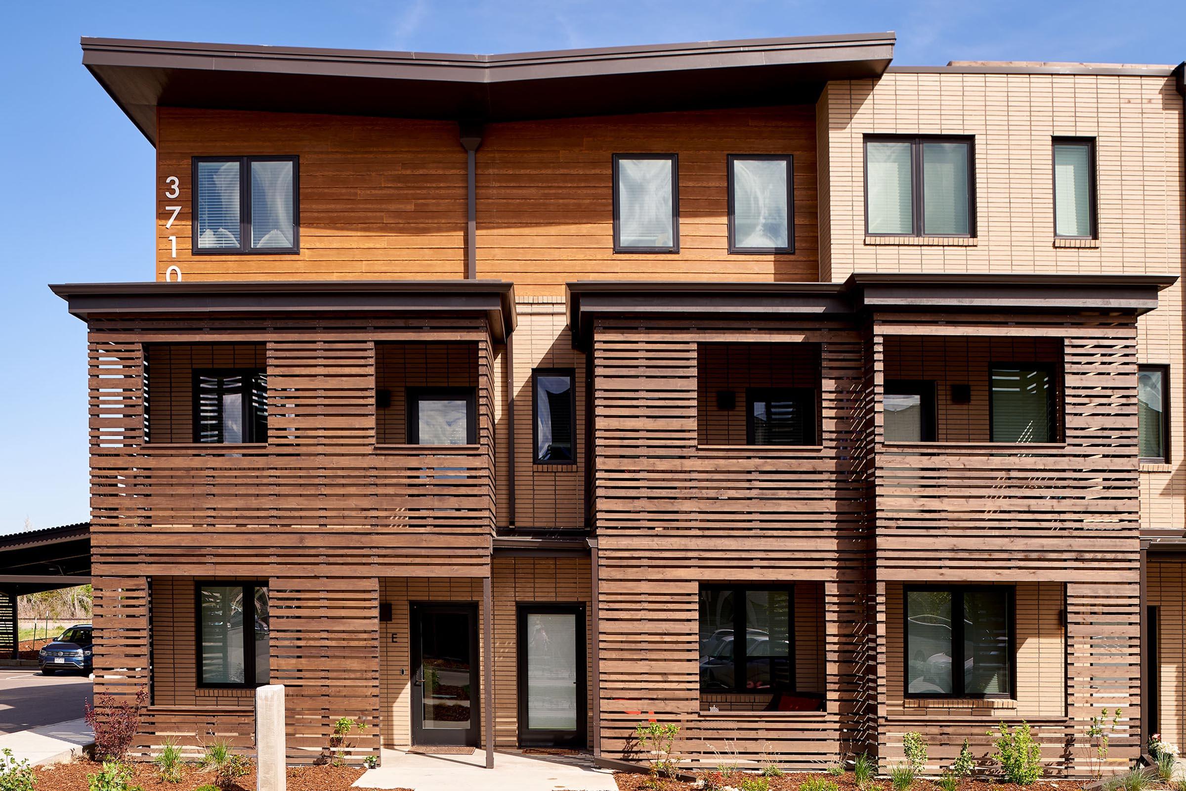 A modern three-story building featuring a combination of wood and brick exteriors. The facade includes horizontal wooden slats, large windows, and a stylish roof design. The entrance is framed by landscaped greenery, creating a welcoming atmosphere. Clear blue skies are visible in the background.
