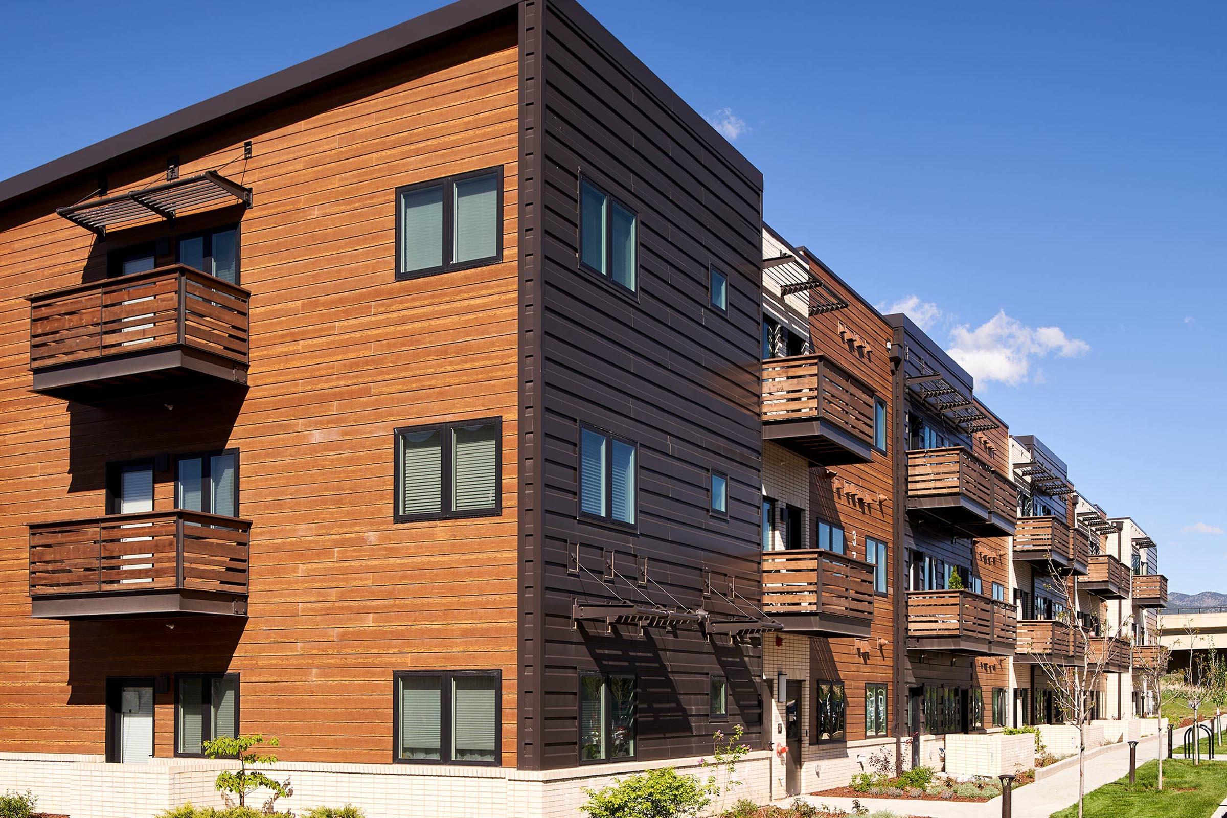 Modern multi-story apartment building featuring wooden exterior, large windows, and balconies. The structure has a contemporary design against a clear blue sky, with landscaped greenery in the foreground.