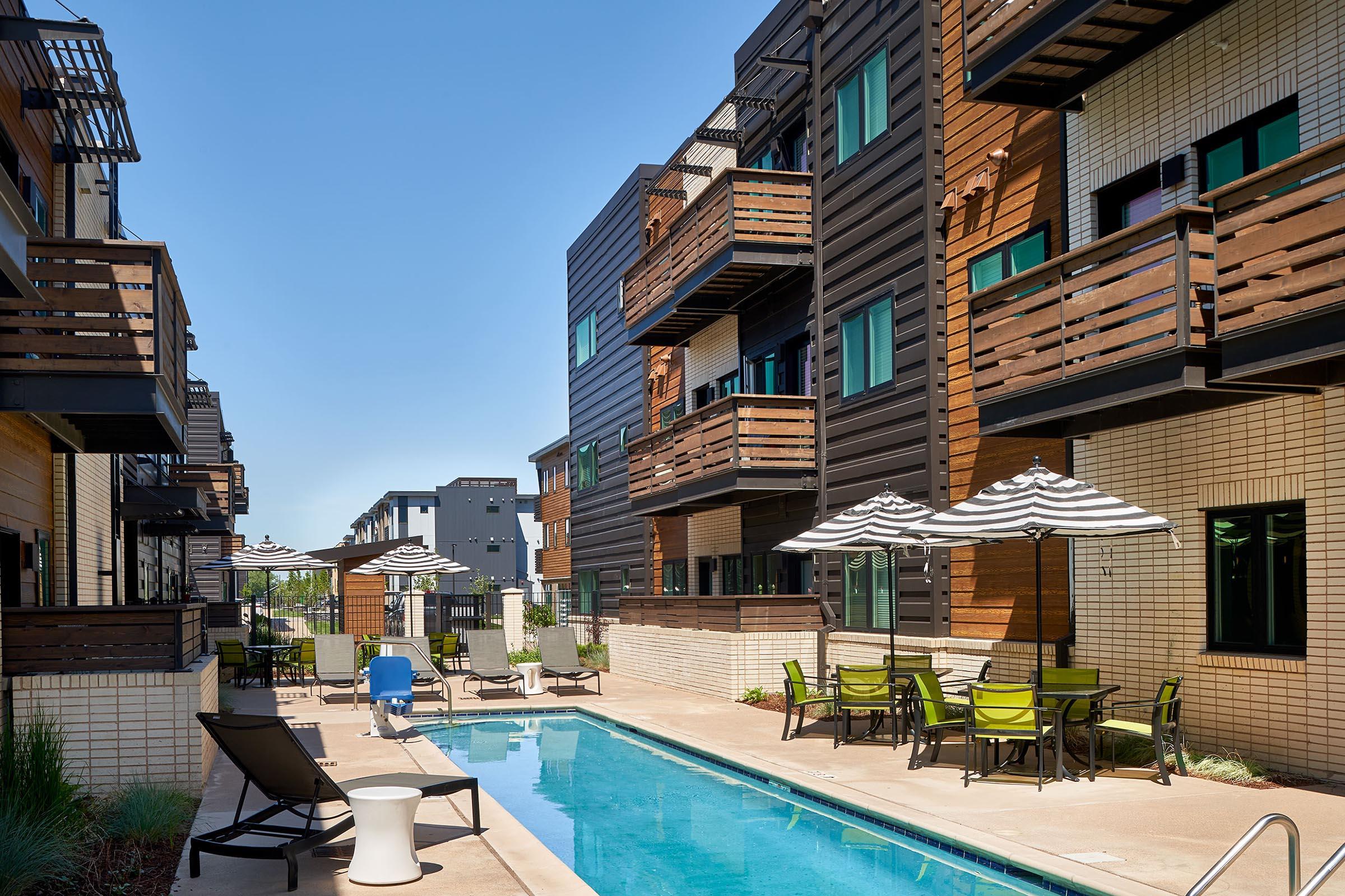 A sunny outdoor courtyard featuring a swimming pool surrounded by lounge chairs and umbrellas. Modern apartment buildings with wooden and brick exteriors line the sides, creating a vibrant and inviting atmosphere for relaxation and leisure.