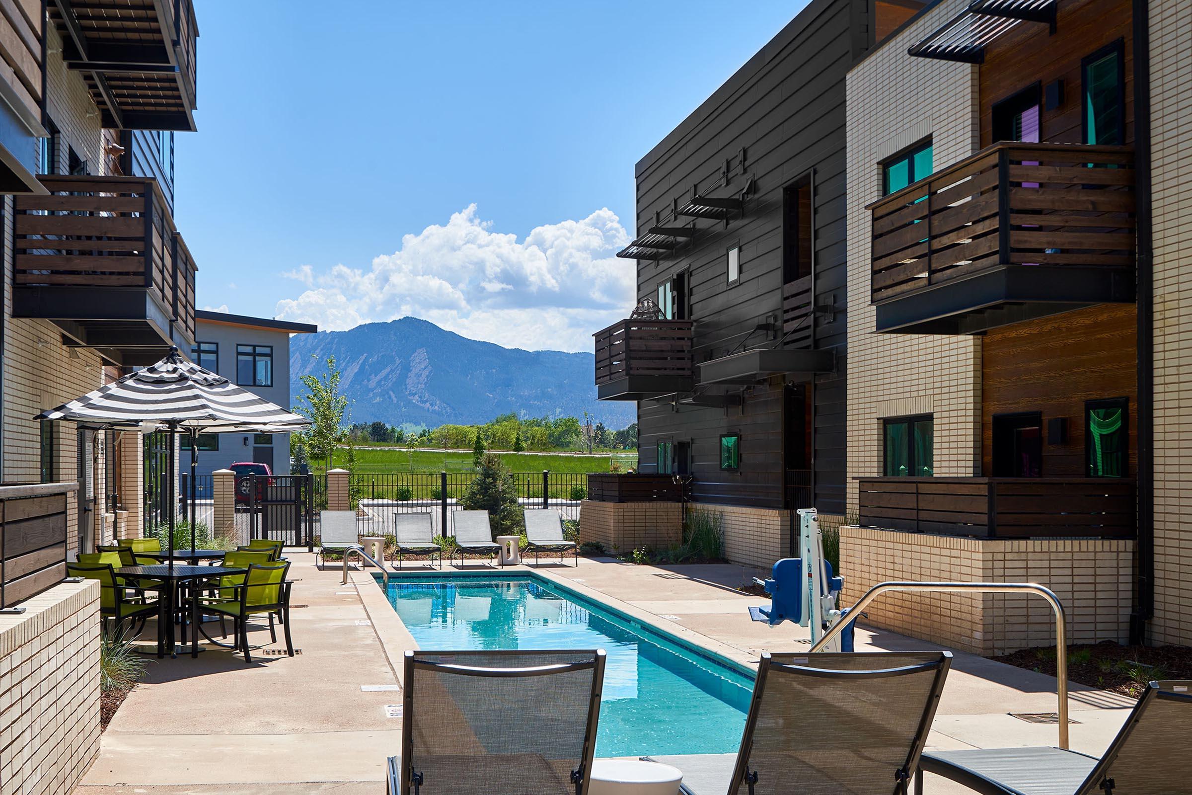 A serene pool area surrounded by modern apartment buildings, with lounge chairs positioned for relaxation. In the background, a stunning mountain range is visible under a clear blue sky, and vibrant green grass adds to the picturesque setting. Shade is provided by an umbrella over a nearby dining table.