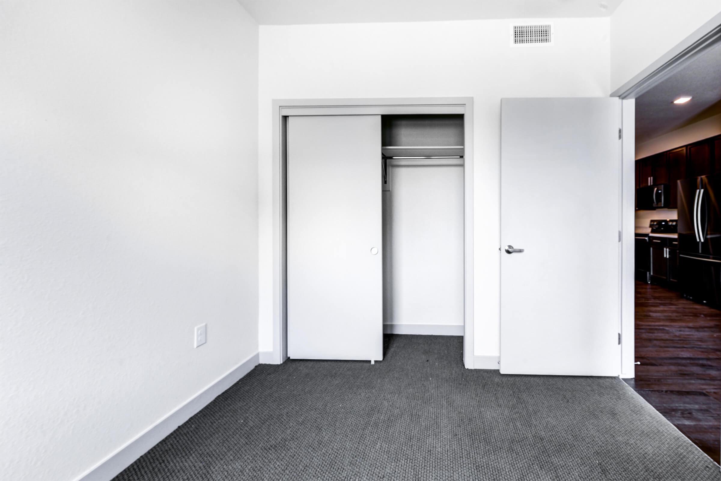 A view of a minimalistic room featuring a closet with closed sliding doors. The walls are white, and there is grey carpet on the floor. A doorway leads to a kitchen area visible in the background, showcasing dark cabinetry and appliances. The room has a bright, clean aesthetic.