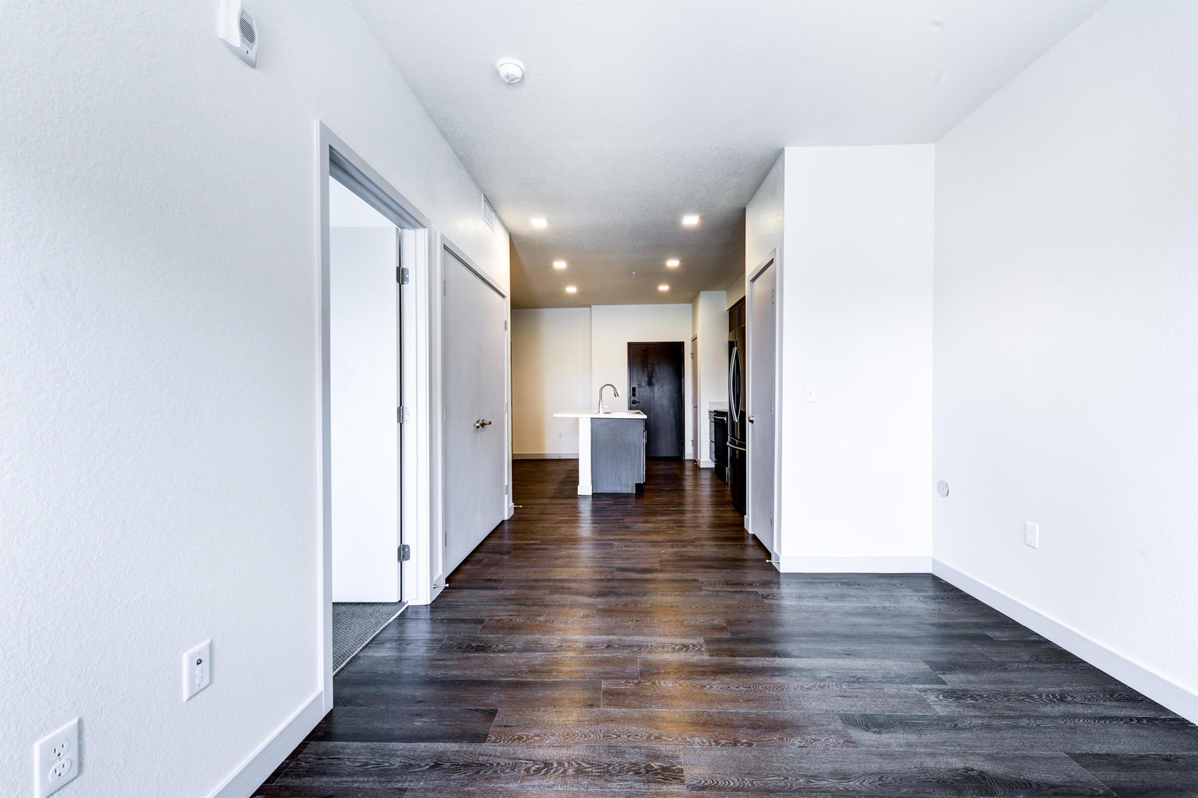 A spacious apartment interior featuring a wooden floor, light-colored walls, and recessed lighting. The view shows an open layout with a kitchen island visible in the background, along with a doorway leading to another room. The overall design is modern and minimalistic.