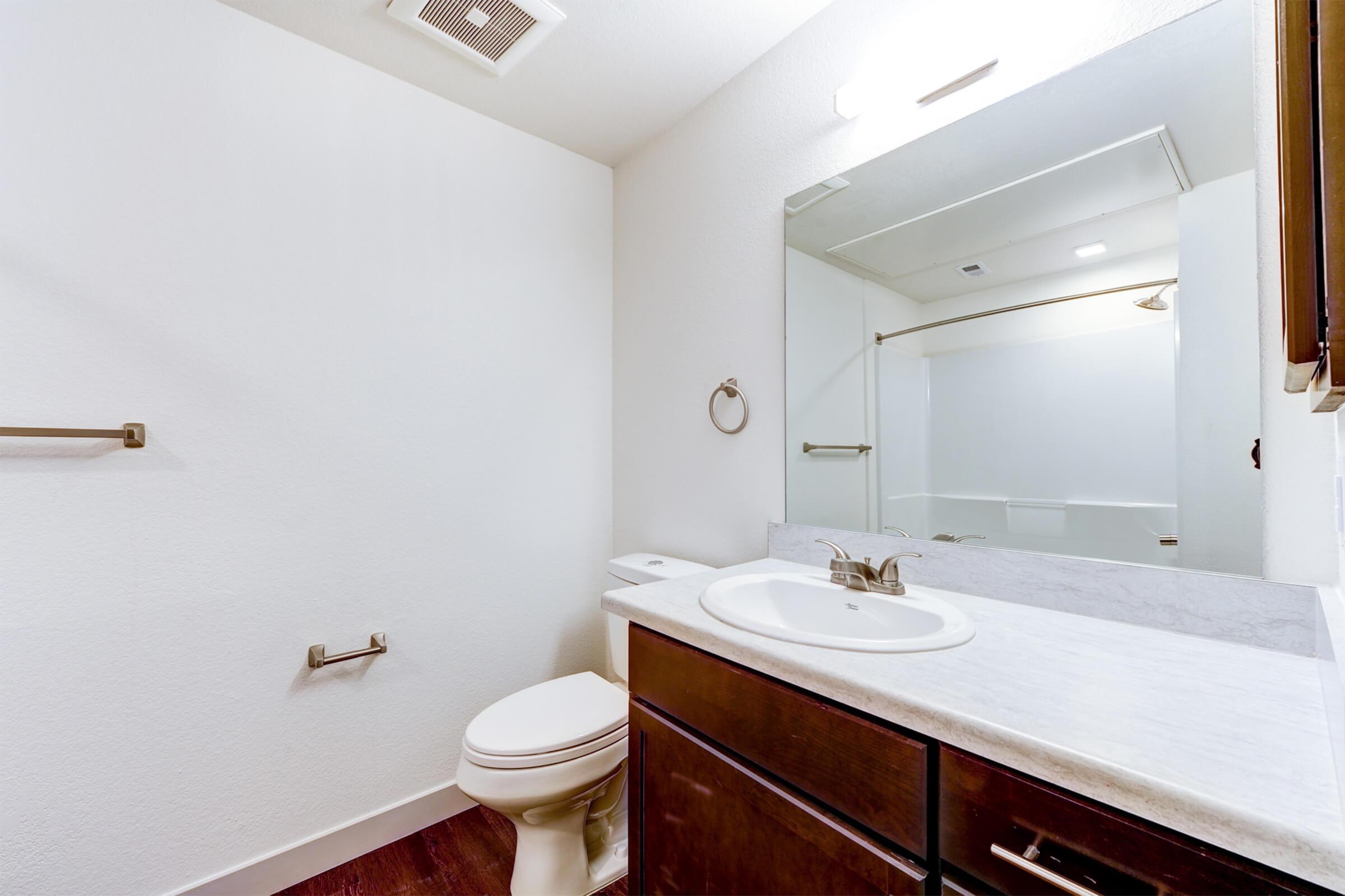 Interior view of a bathroom featuring a white toilet, a marble countertop with a sink, and a large mirror. The walls are painted in a light color, and there is a towel bar on the wall. A shower stall is visible in the background, illuminated by overhead lighting. The floor has dark wood accents.