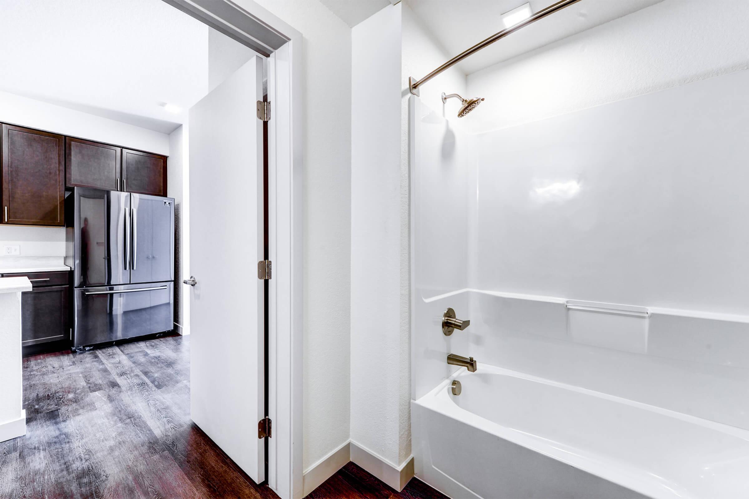 Interior view of a bathroom featuring a white bathtub and shower combination. An open door leads to a modern kitchen with dark wood cabinetry and a stainless steel refrigerator visible in the background. The floor has a dark wood finish, contributing to a contemporary aesthetic. Bright, neutral walls enhance the space.