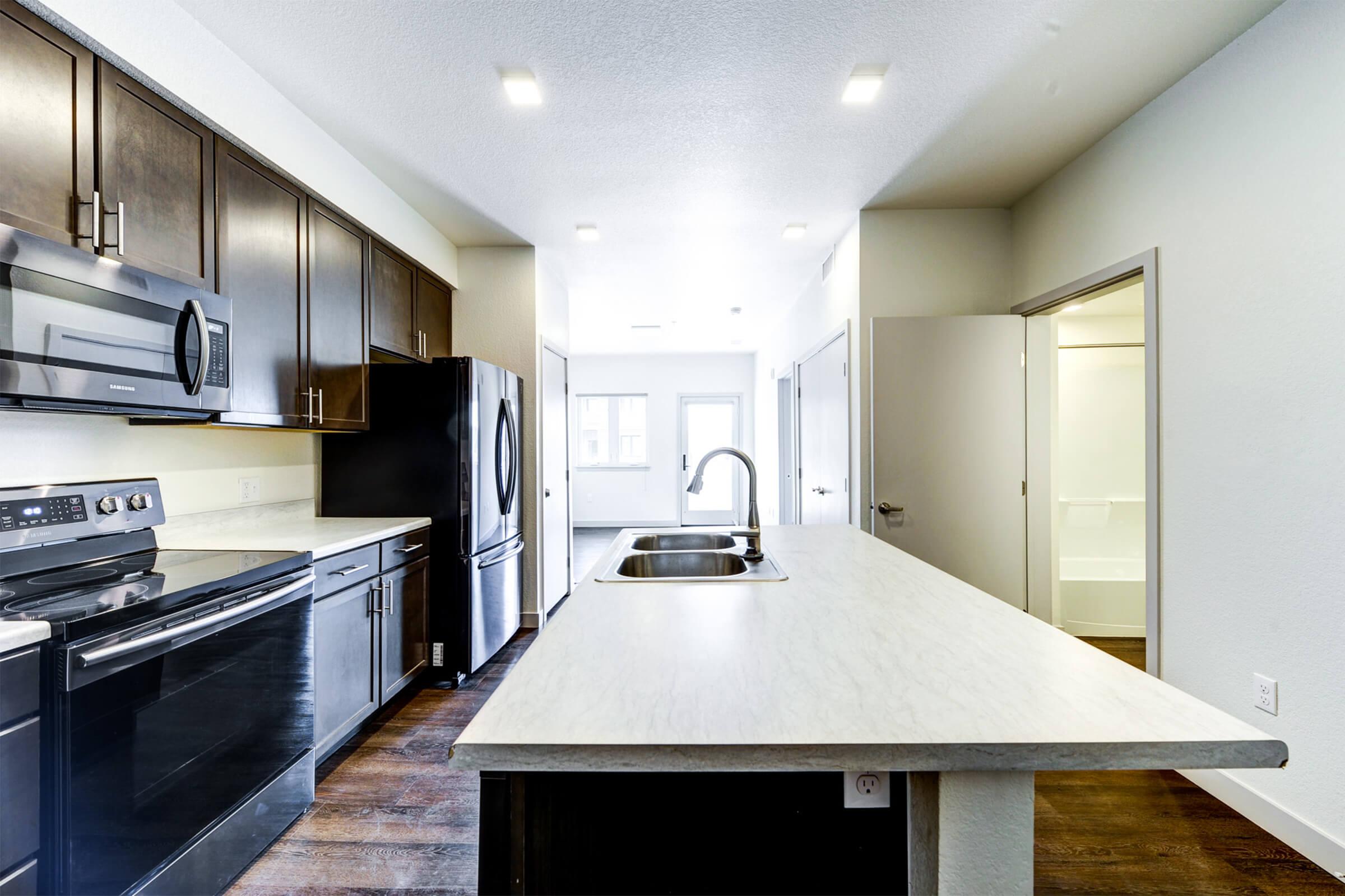 Modern kitchen featuring dark wood cabinets, stainless steel appliances, a large island with a sink, and an open layout. Bright lighting illuminates the space, and there are light-colored walls. A doorway leads to another room, suggesting an airy and functional design.