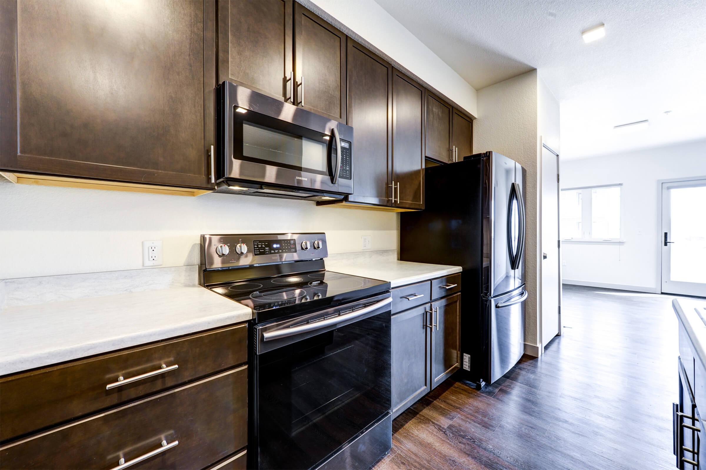 Modern kitchen featuring dark wood cabinets, stainless steel appliances including a microwave and oven, a black refrigerator, and a countertop with a light-colored surface. The kitchen has hardwood flooring and a doorway leading to a well-lit area, suggesting an open-concept design.
