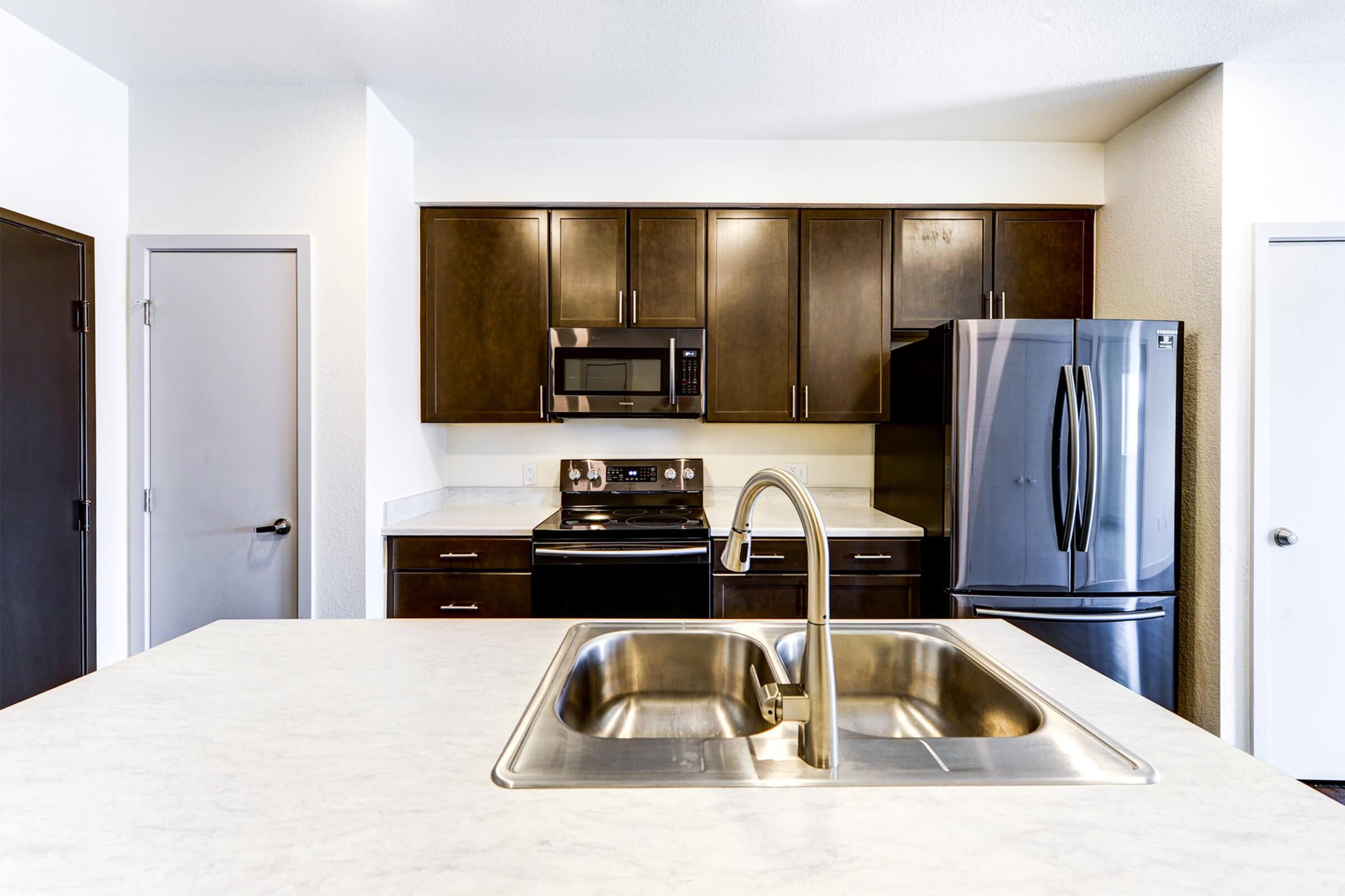 A modern kitchen featuring dark wood cabinets, stainless steel appliances including a refrigerator and microwave, and a double sink with a chrome faucet. The countertop is made of light-colored stone, providing a contrast to the cabinetry. There are doors leading to other areas of the space.