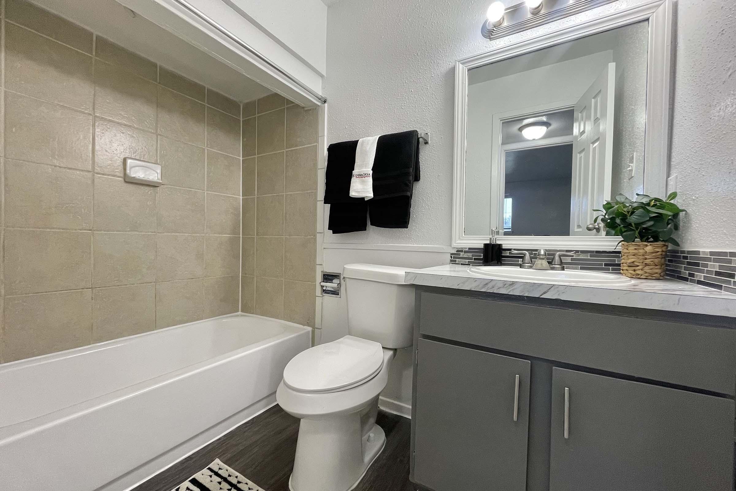 A modern bathroom featuring a bathtub with a beige tiled surround, a white toilet, and a grey vanity with a marble countertop. The mirror above the sink is framed, and there are black towels hung on a rack. A small potted plant decorates the vanity, and a textured rug lies on the floor.