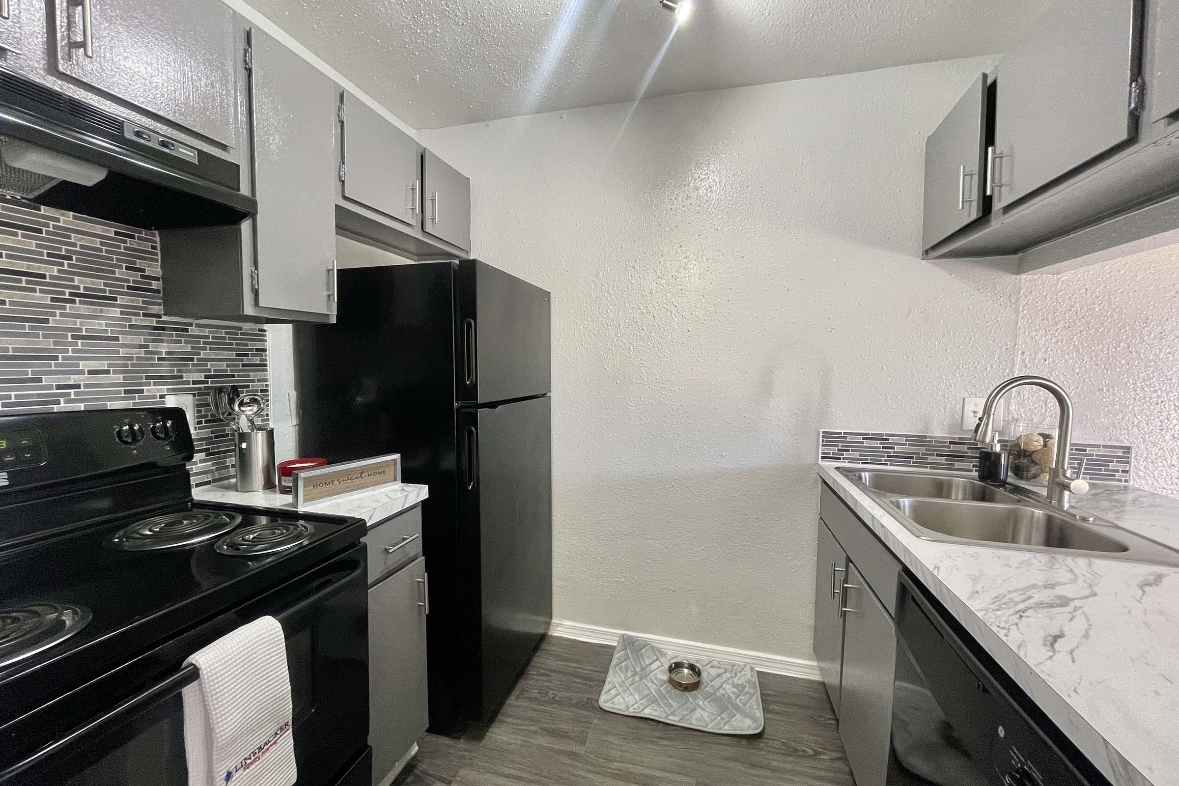 Modern kitchen featuring gray cabinets, a black refrigerator, and a black stove. The countertop is made of white marble with a stainless steel sink. The backsplash has decorative tiles. A dog bowl is on the floor, and the walls are painted white. Brightly lit with overhead lights.
