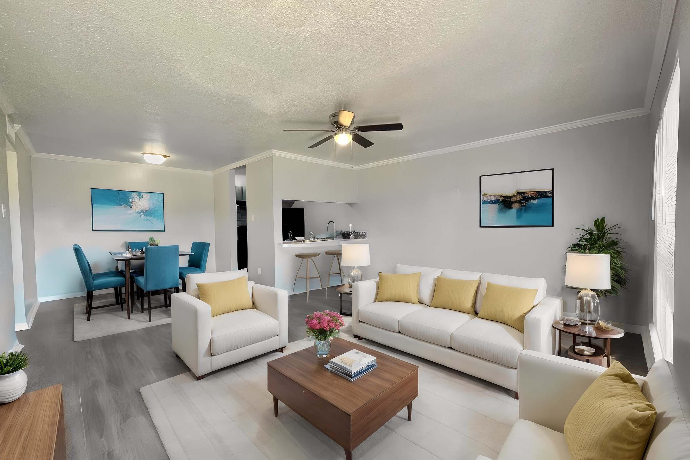 A modern living room featuring a white sofa with yellow pillows, a wooden coffee table, and a plant. In the background, there is a dining area with a round table and blue chairs, along with a kitchen visible. The walls are light gray, and there are two framed artworks on display.