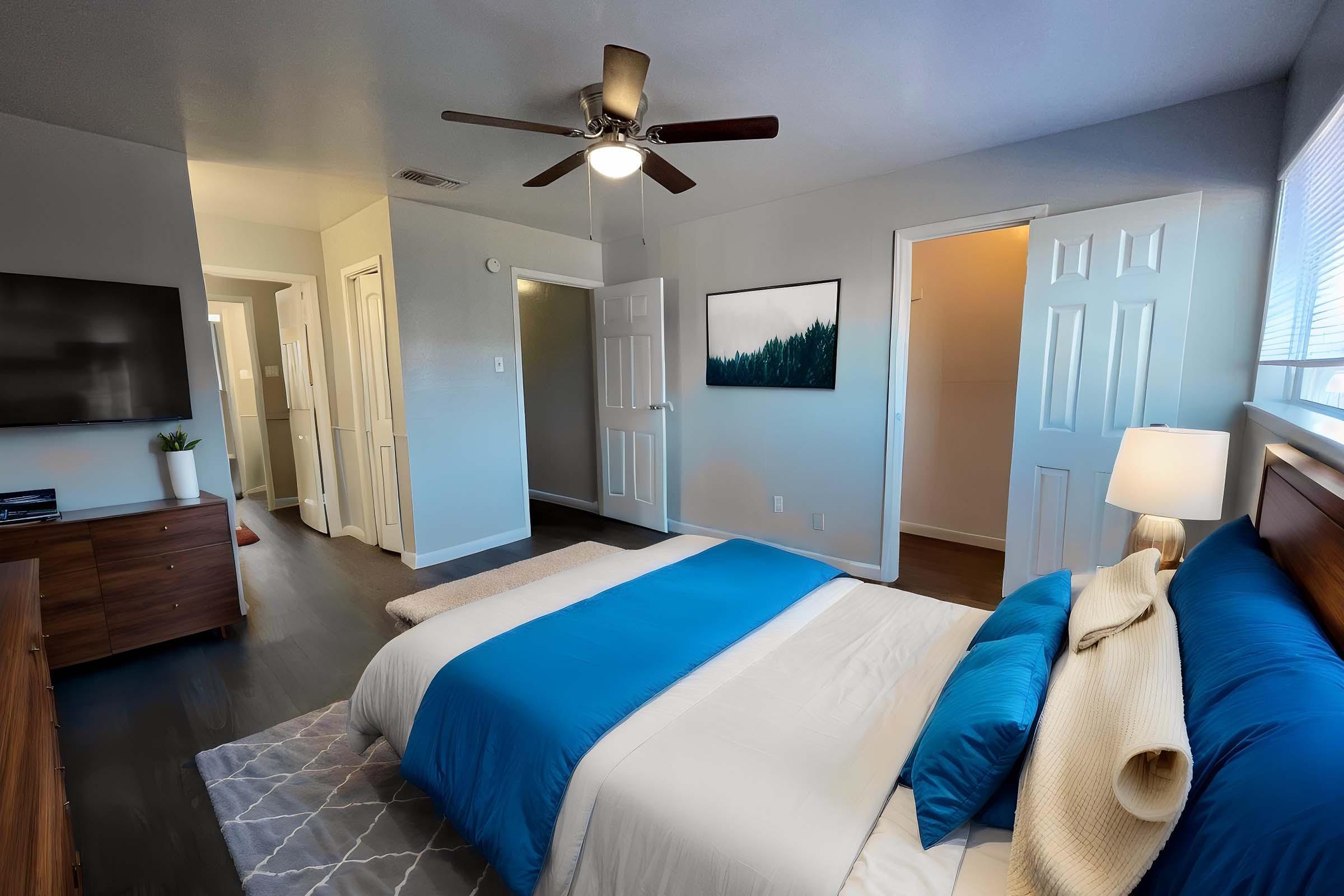 A well-lit bedroom featuring a queen-sized bed with a blue and white bedspread, a wooden nightstand, and a ceiling fan. A modern art piece hangs on the wall, and there are two doors leading to other rooms. The floor is dark wood, and a soft rug is placed beside the bed.