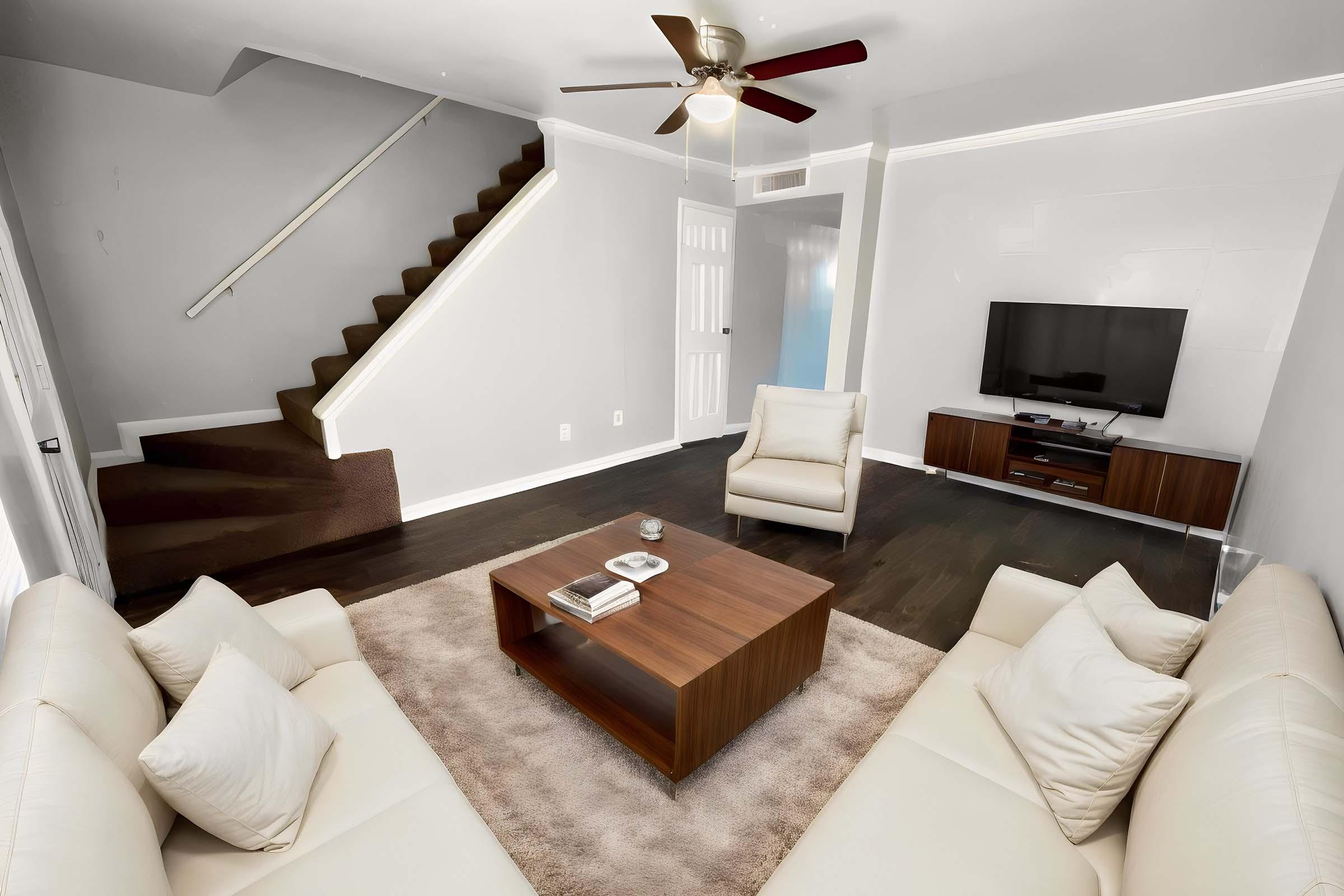 A modern living room featuring a white leather sofa set and a wooden coffee table on a light rug. There is a staircase with brown steps in the background, a flat-screen TV mounted on the wall, and a single armchair. The walls are painted light gray, creating a bright and spacious atmosphere.