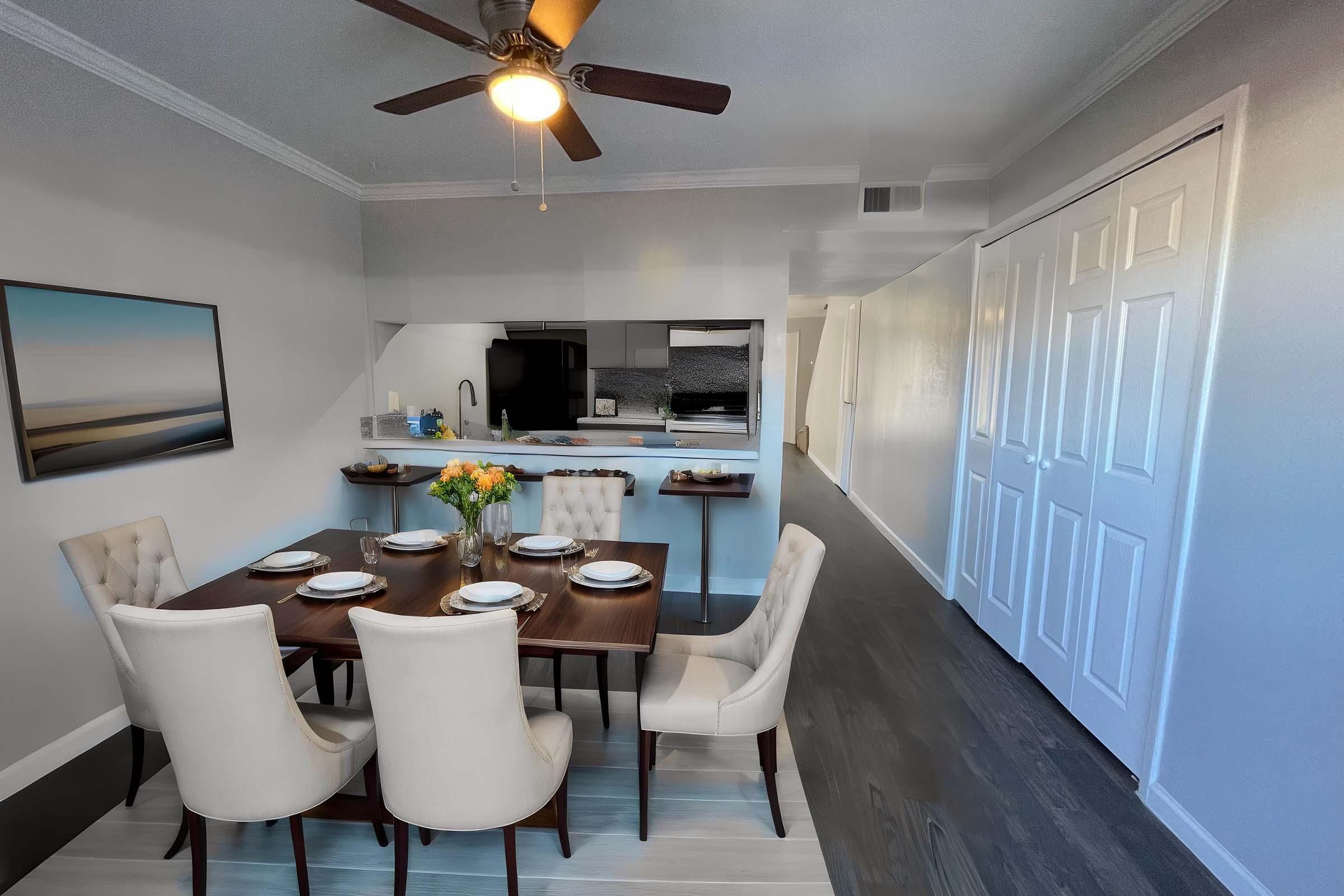 A modern dining area featuring a wooden table set for six with white chairs. A bouquet of flowers sits in the center of the table. The background includes a kitchen with sleek appliances, a ceiling fan, and a light-colored wall. A painting of a serene landscape hangs on the wall.