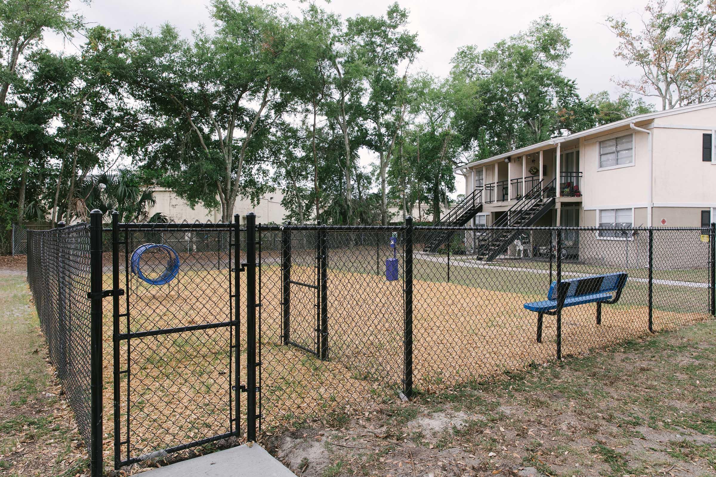 A fenced dog park with a sandy area, featuring a blue bench and a circular disc attached to the fence. Surrounding trees provide shade, and an apartment building is visible in the background. The entrance gate is open, inviting pets and their owners to enjoy the space.