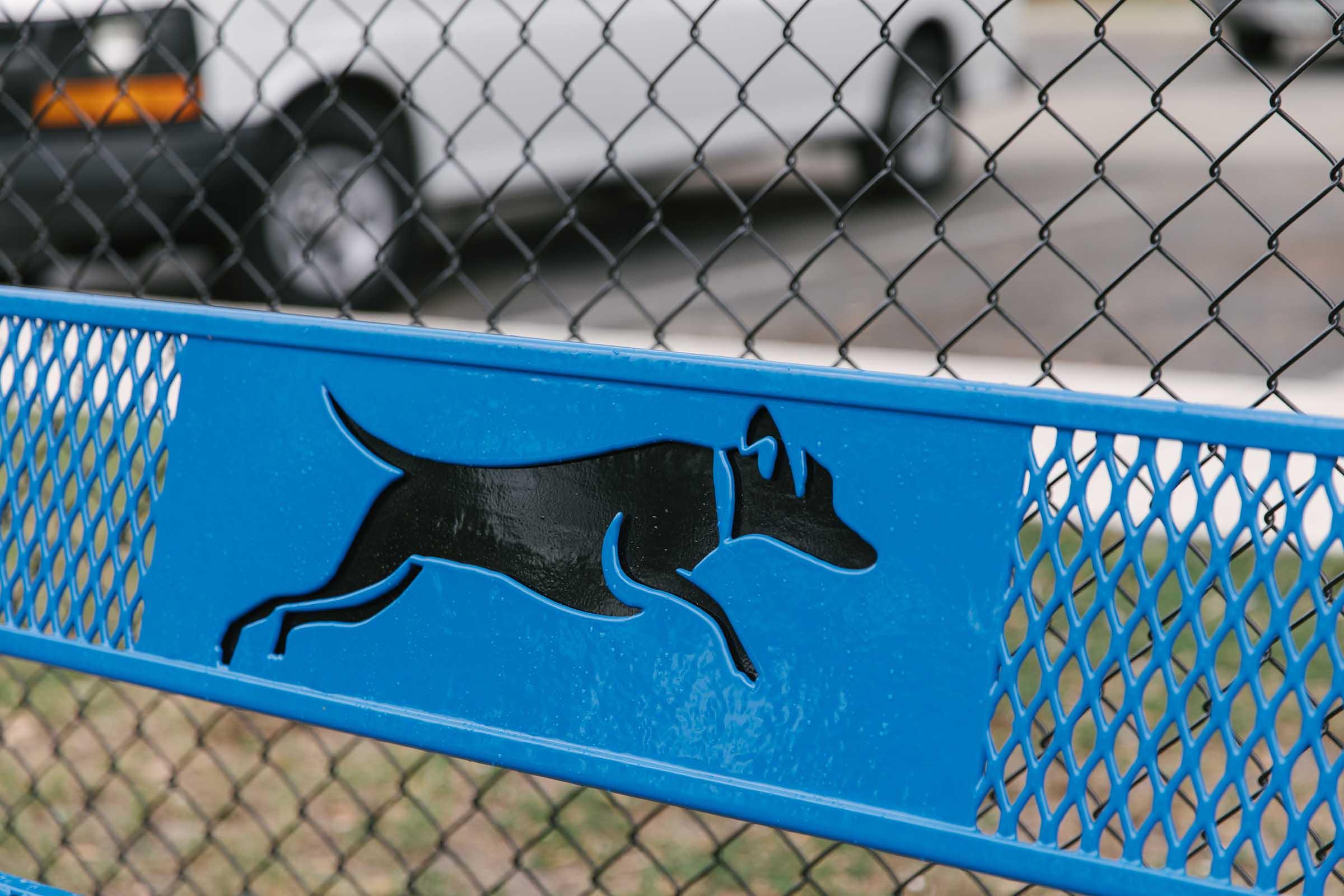 A blue bench with a silhouette of a running dog cut out in the backrest, set against a chain-link fence. A car is parked in the background, and the ground is grassy.