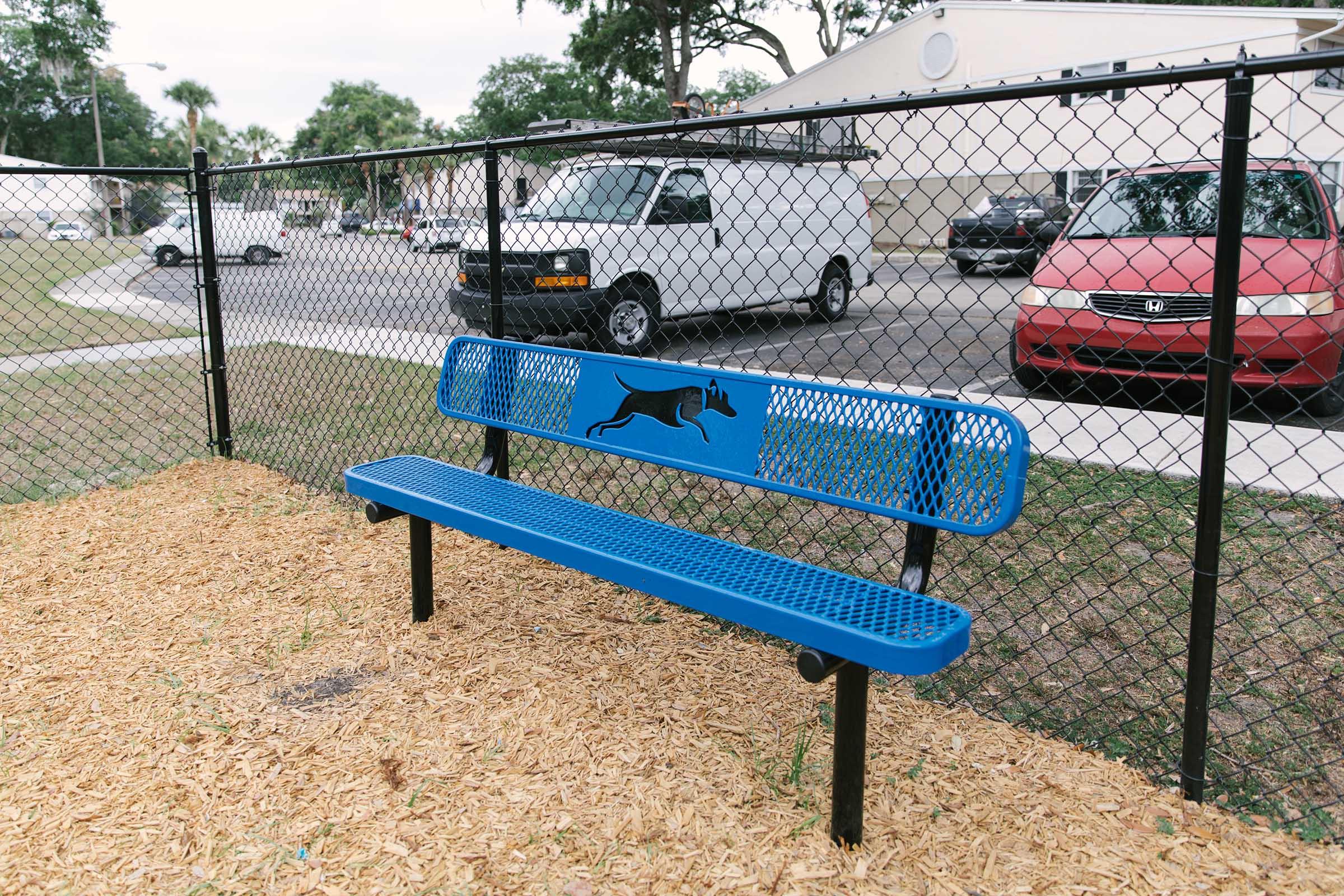A blue bench with a dog silhouette design, located in a fenced area covered with wood chips. In the background, there are parked vehicles and a building. The setting appears to be a park or pet-friendly area.