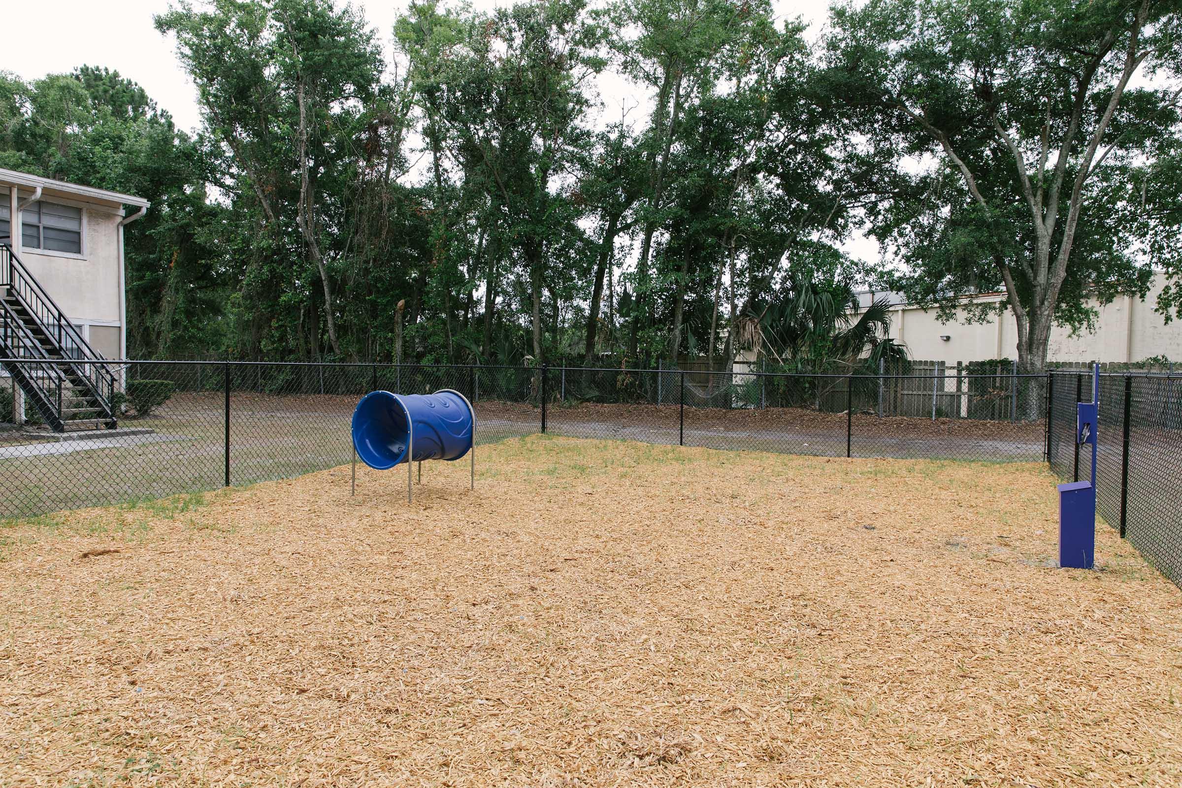 A fenced playground area featuring wood chips as ground cover, with a blue tunnel-shaped slide and grassy edges. In the background, there are trees and a building, creating a natural setting. The playground is empty, suggesting it's a quiet, open space for children to play.