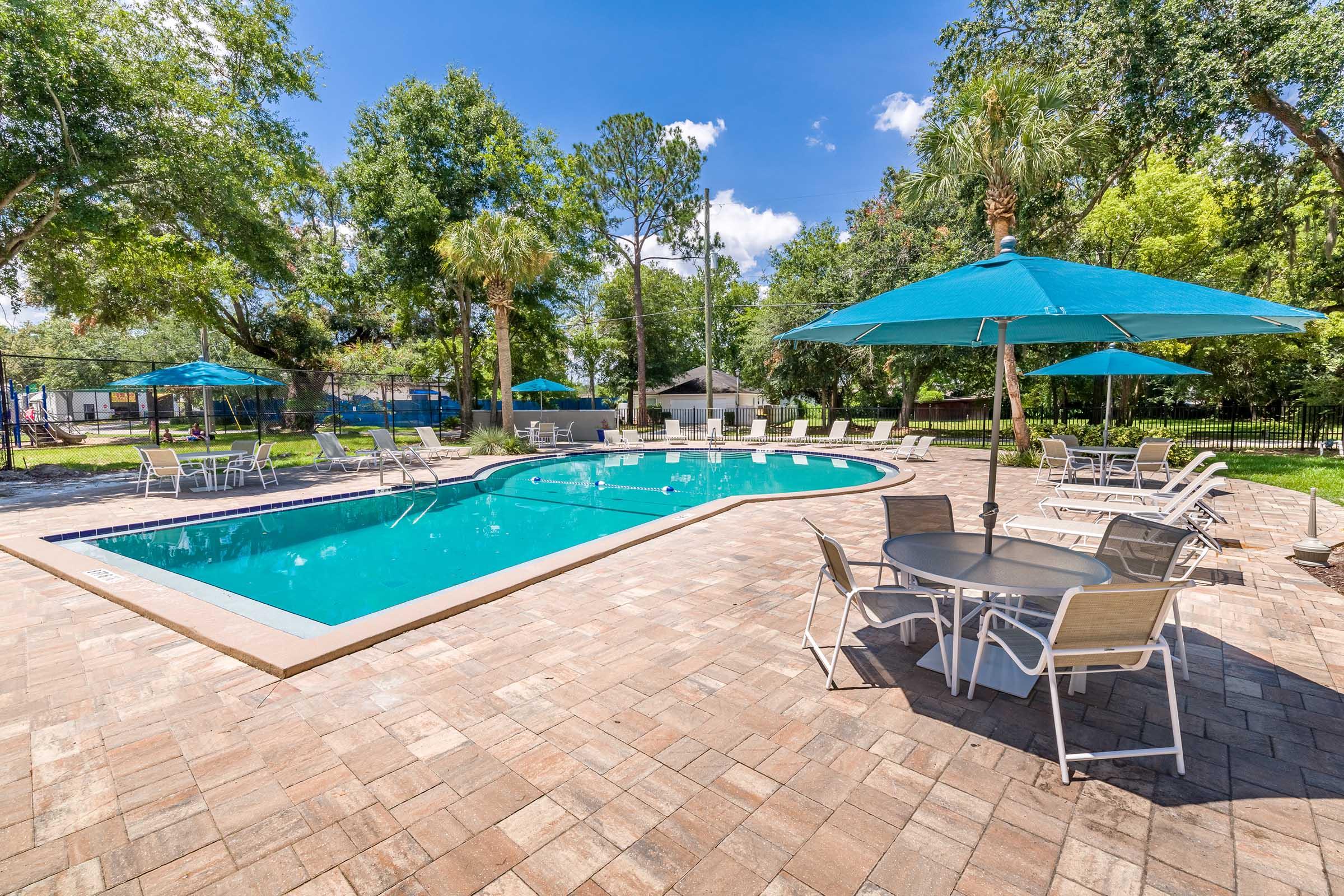 A sunny outdoor pool area featuring a clear blue swimming pool surrounded by lounge chairs and tables. Bright turquoise umbrellas provide shade, with lush greenery and trees in the background, creating a relaxing and inviting atmosphere perfect for leisure and social gathering.
