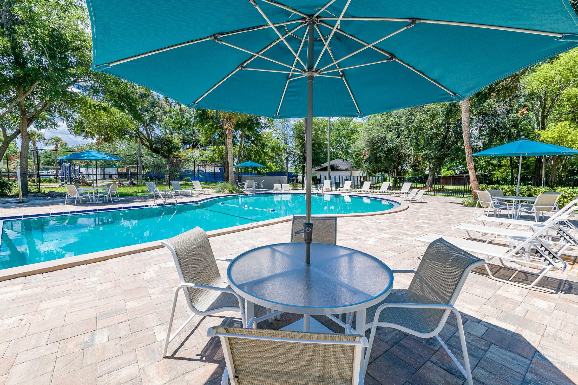 A vibrant swimming pool scene featuring clear blue water surrounded by patio tiles. Several lounge chairs and tables with turquoise umbrellas provide shaded seating. Lush green trees and a grassy area create a relaxing atmosphere in a sunny outdoor setting.
