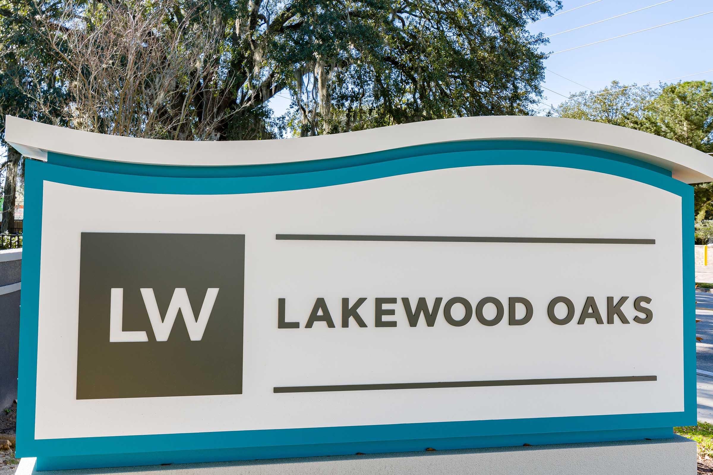Sign for Lakewood Oaks featuring the initials "LW" in a stylized font. The sign is modern and prominently displayed, with a blue and white color scheme. It is set against a backdrop of greenery, suggesting a residential community or neighborhood.