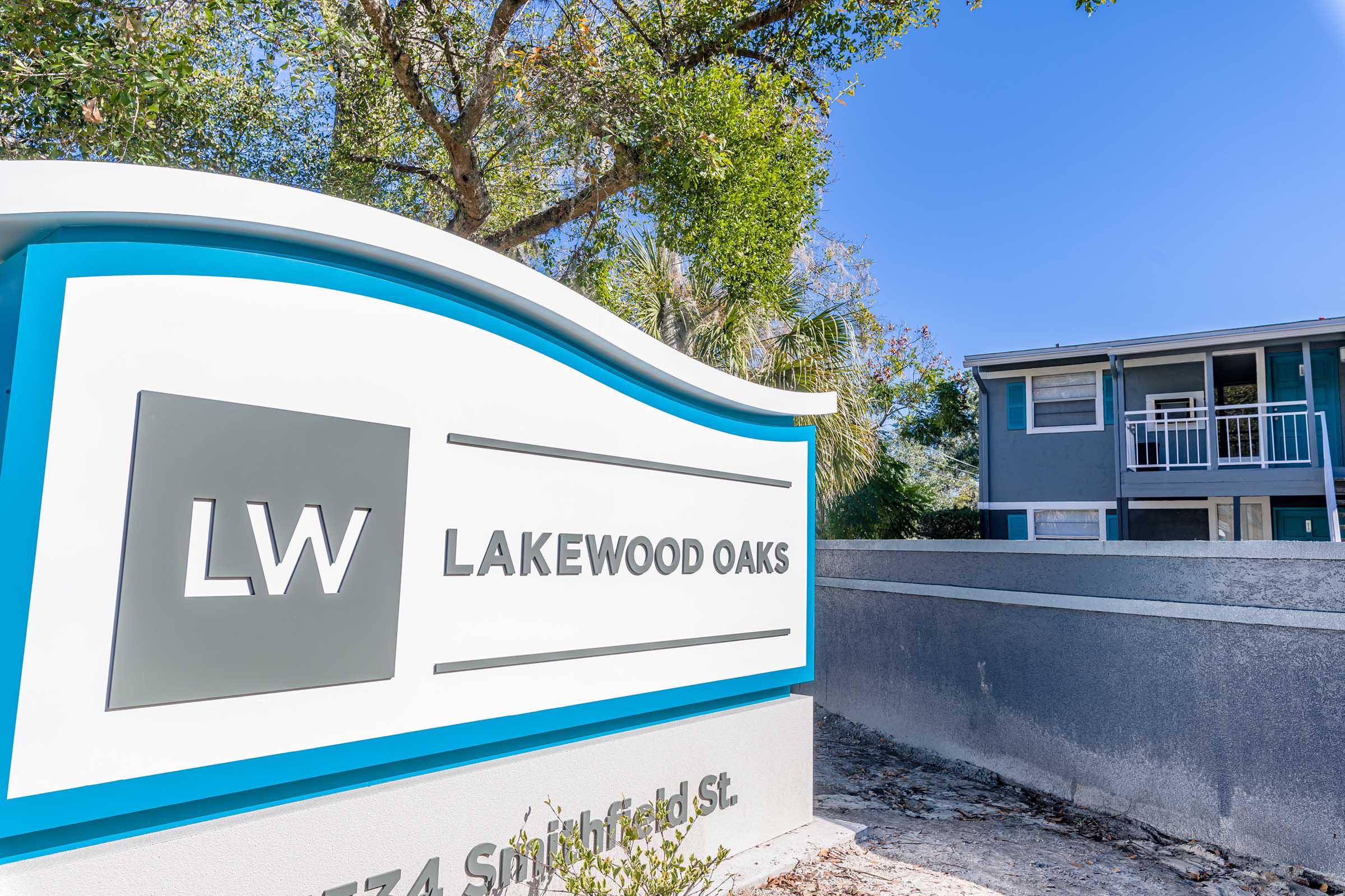 Signage for Lakewood Oaks, featuring the letters "LW" and the address 574 Snipefield St. The sign is modern and prominently displayed, surrounded by greenery, with a building visible in the background.