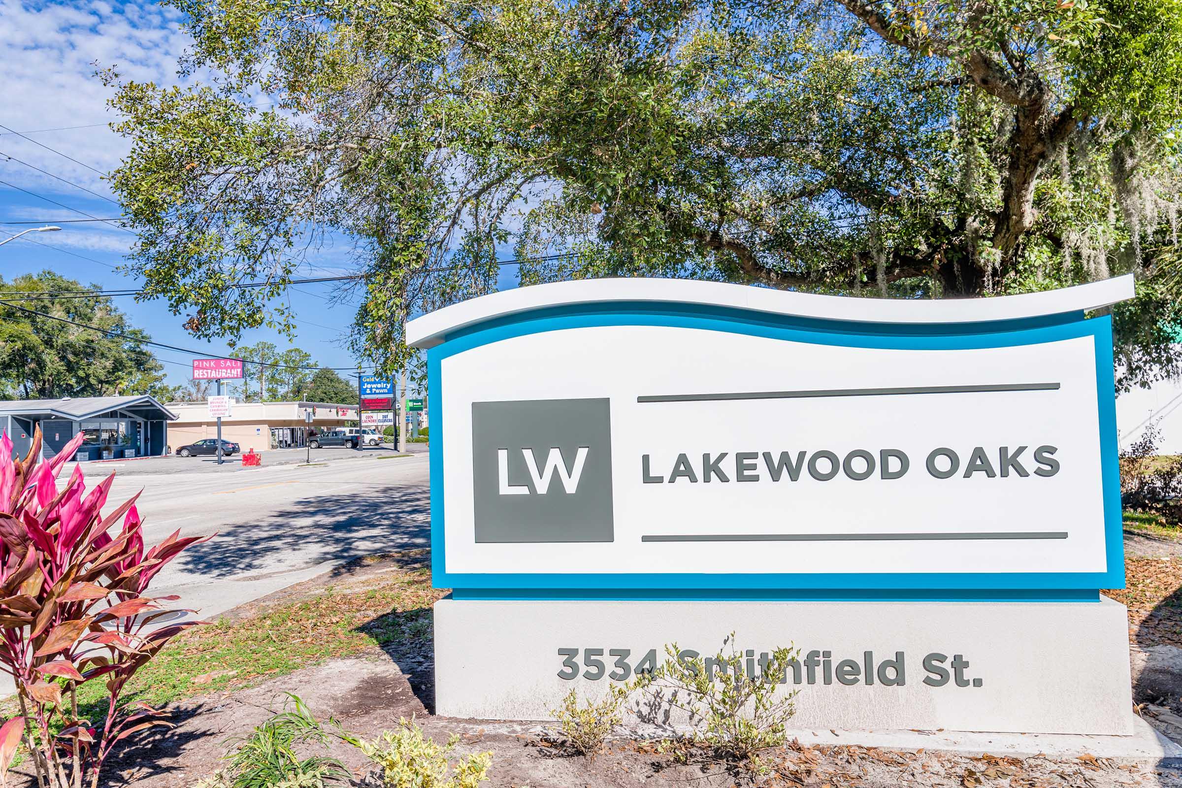 Sign for Lakewood Oaks located at 3534 Springfield St, featuring the initials "LW" prominently. The sign is surrounded by green plants and trees, with businesses visible in the background. The sky is clear, and the scene represents a welcoming community area.