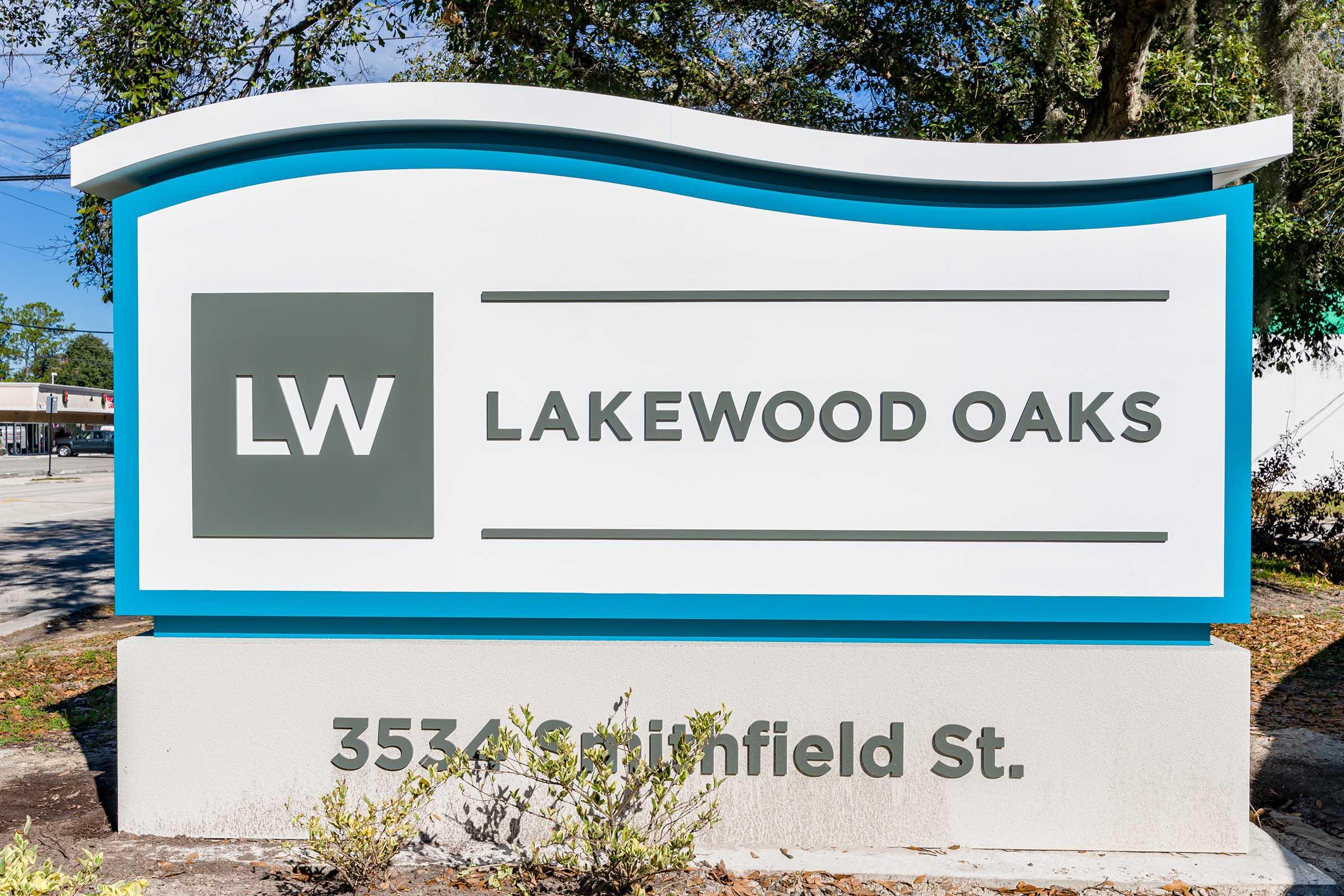 Sign for Lakewood Oaks featuring the letters "LW," with the address "3534 S. Plainfield St." displayed below. The sign has a modern design, featuring a curved top in blue and gray tones, set against a landscaped backdrop.