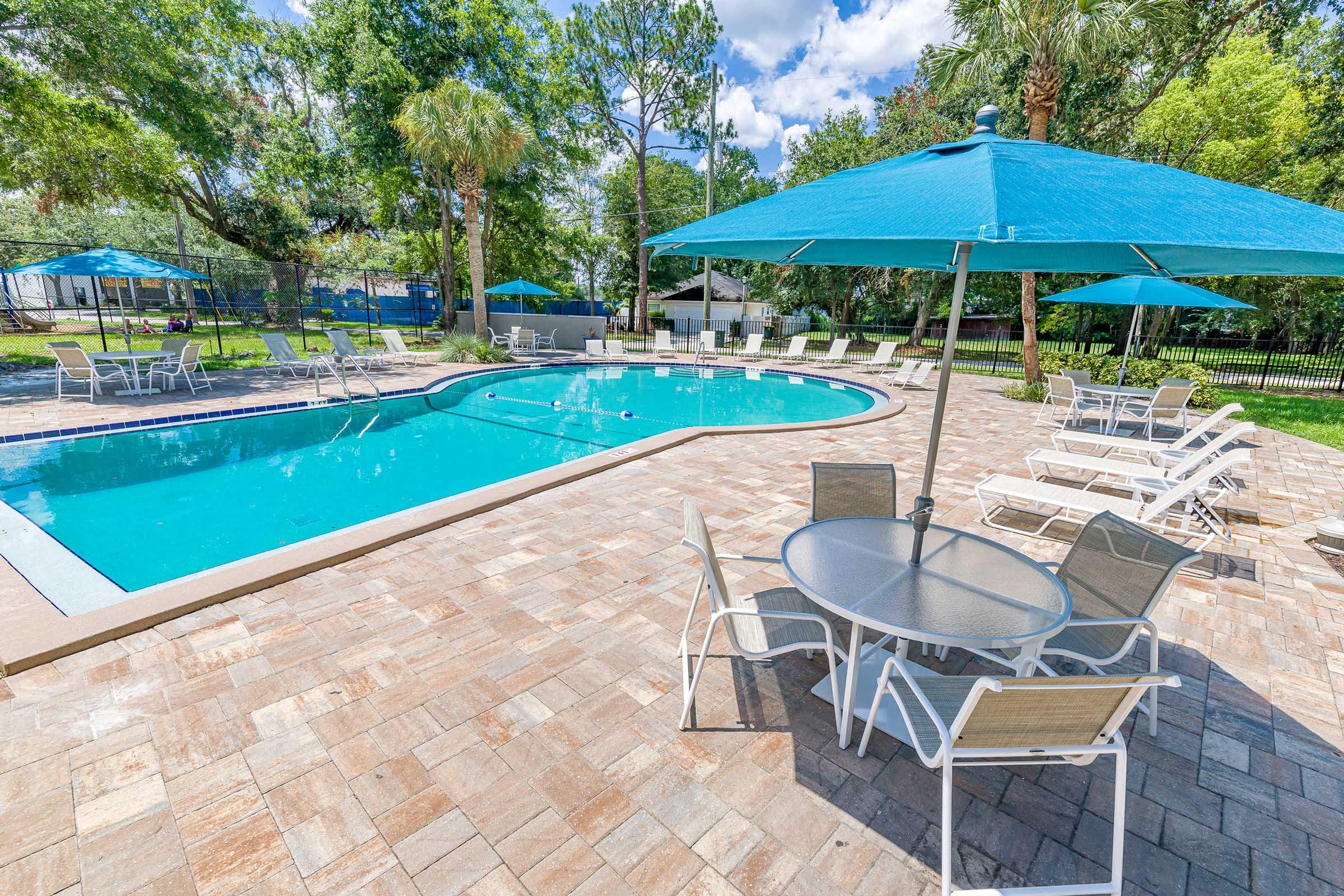 A bright outdoor pool area featuring a clear blue pool surrounded by lounge chairs and tables. Large turquoise umbrellas provide shade, while lush greenery and palm trees create a relaxing atmosphere. The space is well-maintained and inviting for leisure and enjoyment.