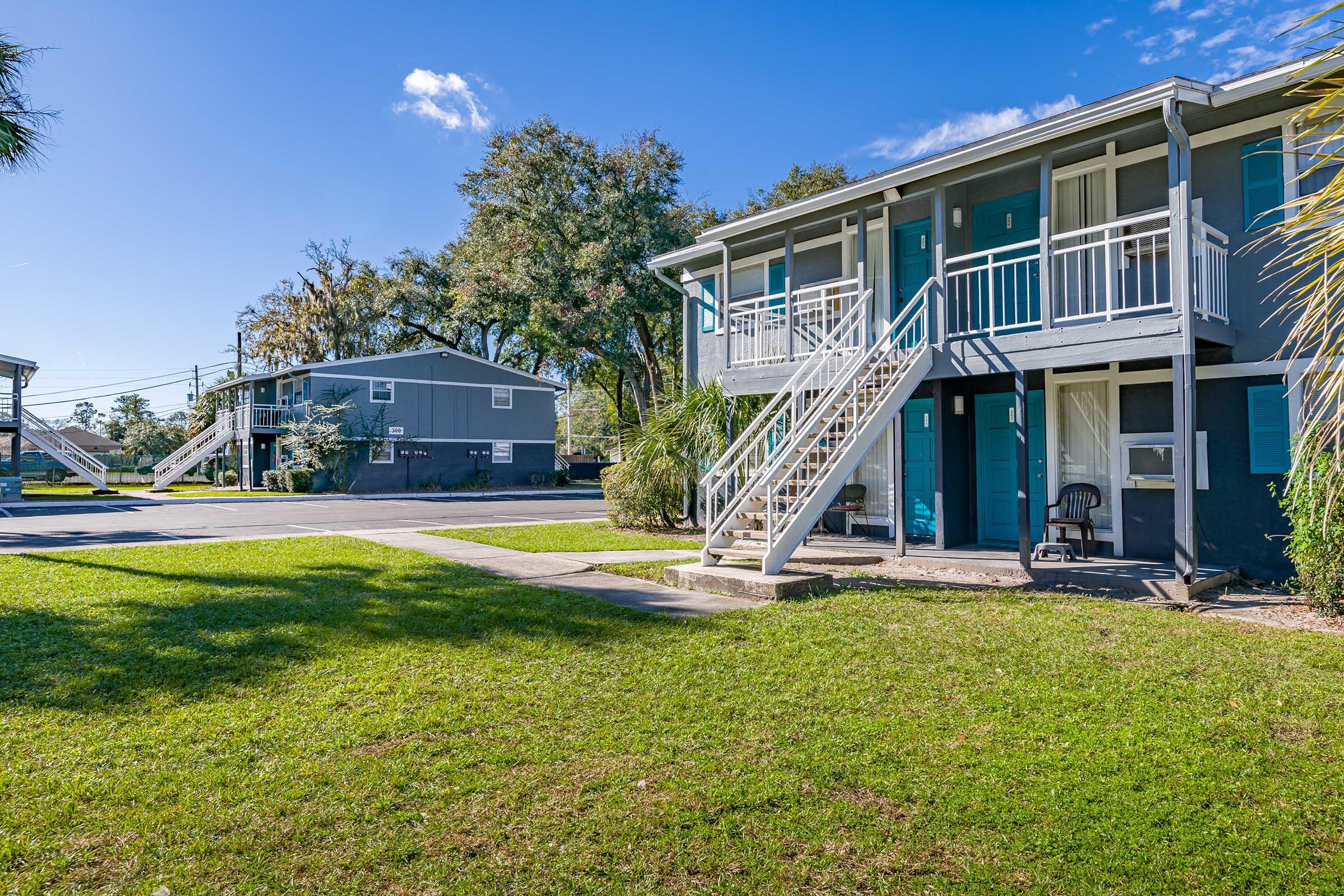 A two-story building with several apartments, featuring light blue doors and white railings. In the foreground, there is a grassy area with a small path. Two trees and palm plants are visible, along with another building in the background. The sky is clear and blue, indicating a sunny day.