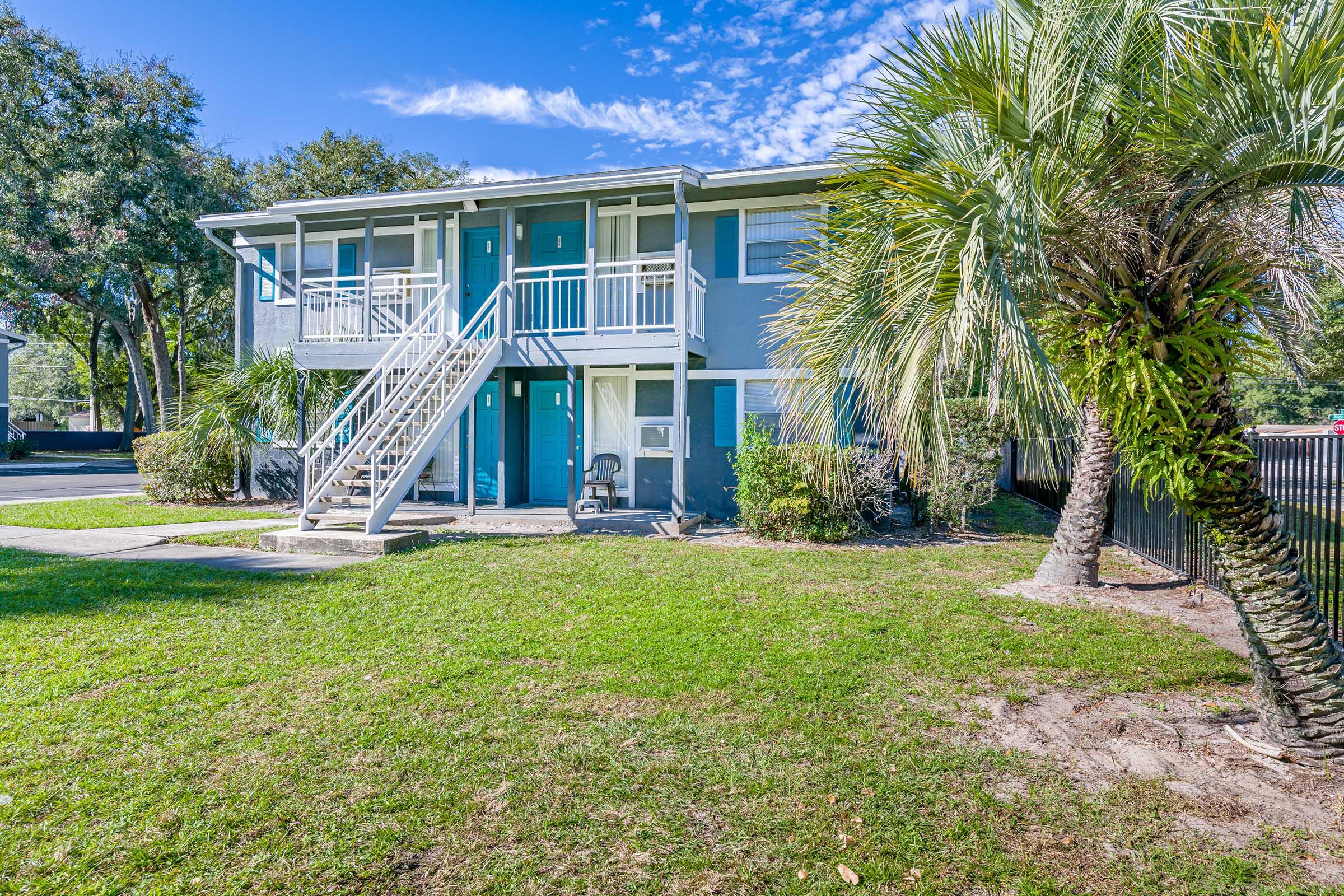 Two-story residential building with light blue exterior and white railings, surrounded by lush greenery and palm trees. A staircase leads to the upper level, and there is a patch of grass in front. The scene is set under a clear blue sky with a few scattered clouds.