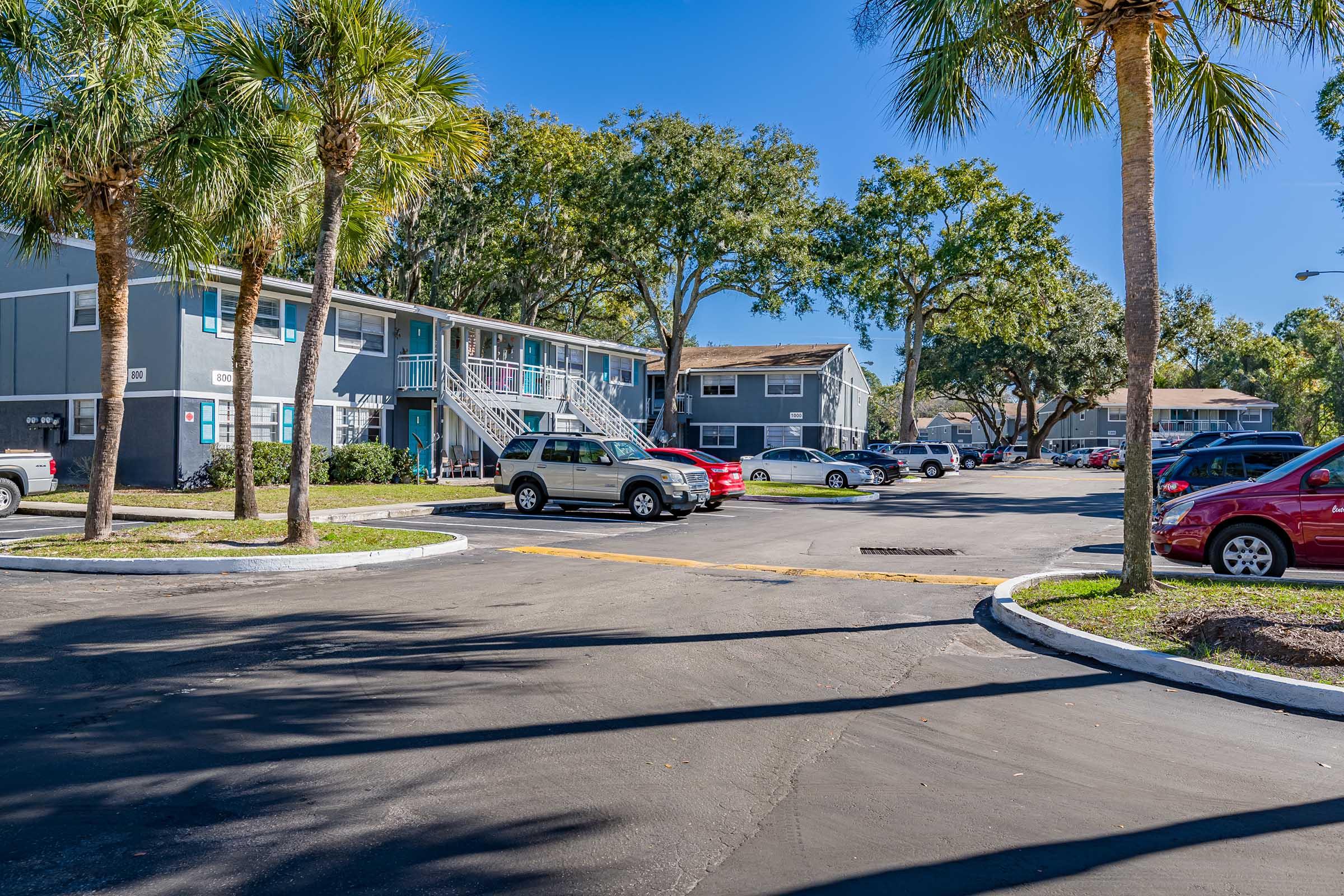 A residential apartment complex with two-story buildings surrounded by palm trees. The foreground shows a parking lot with several cars, and the background features more buildings and greenery under a blue sky. The scene is bright and inviting, typical of a warm climate.
