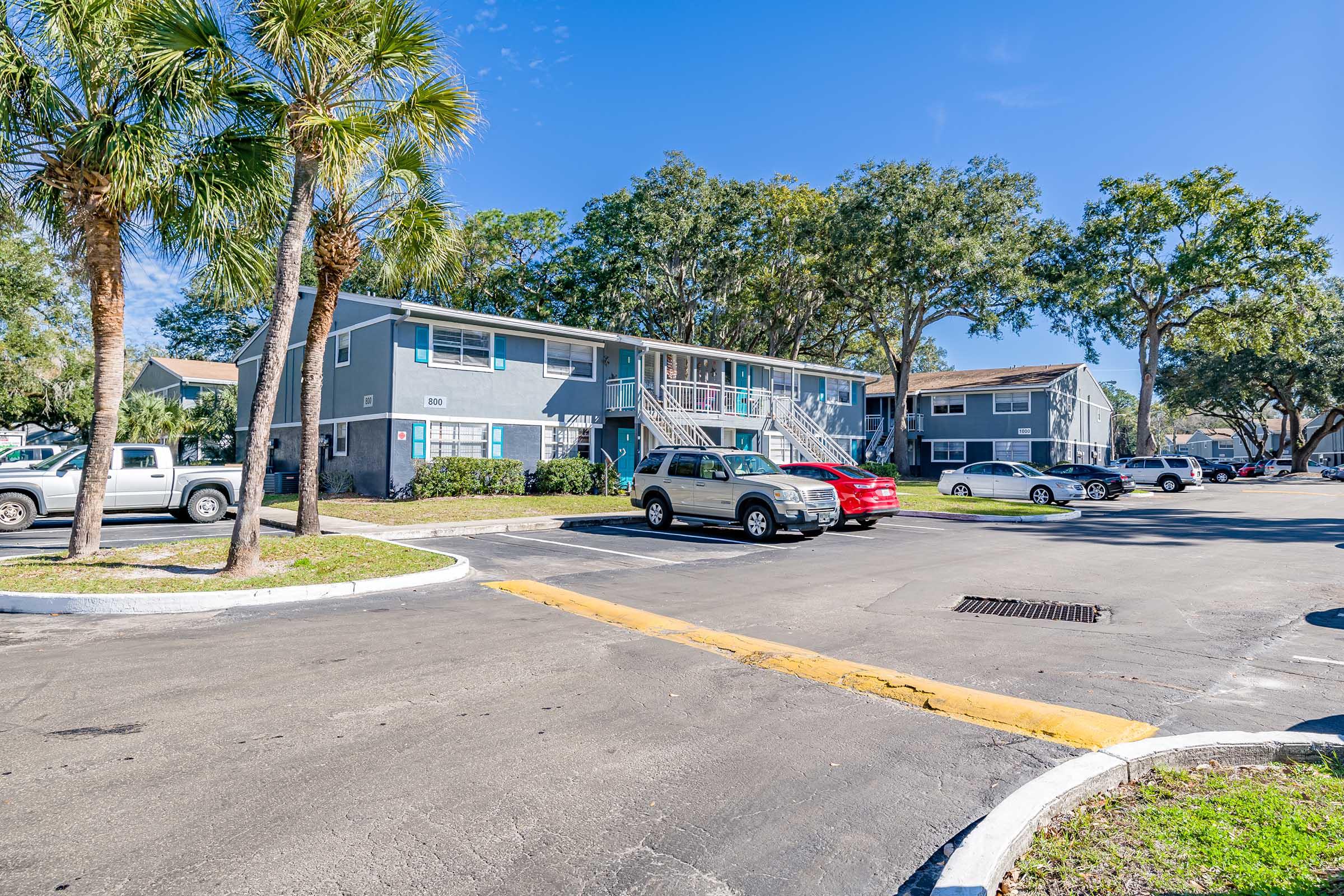 A residential apartment complex featuring two-story buildings surrounded by palm trees and green foliage. Parking spaces are visible in front, with several cars parked. The sky is clear and blue, creating a sunny atmosphere.