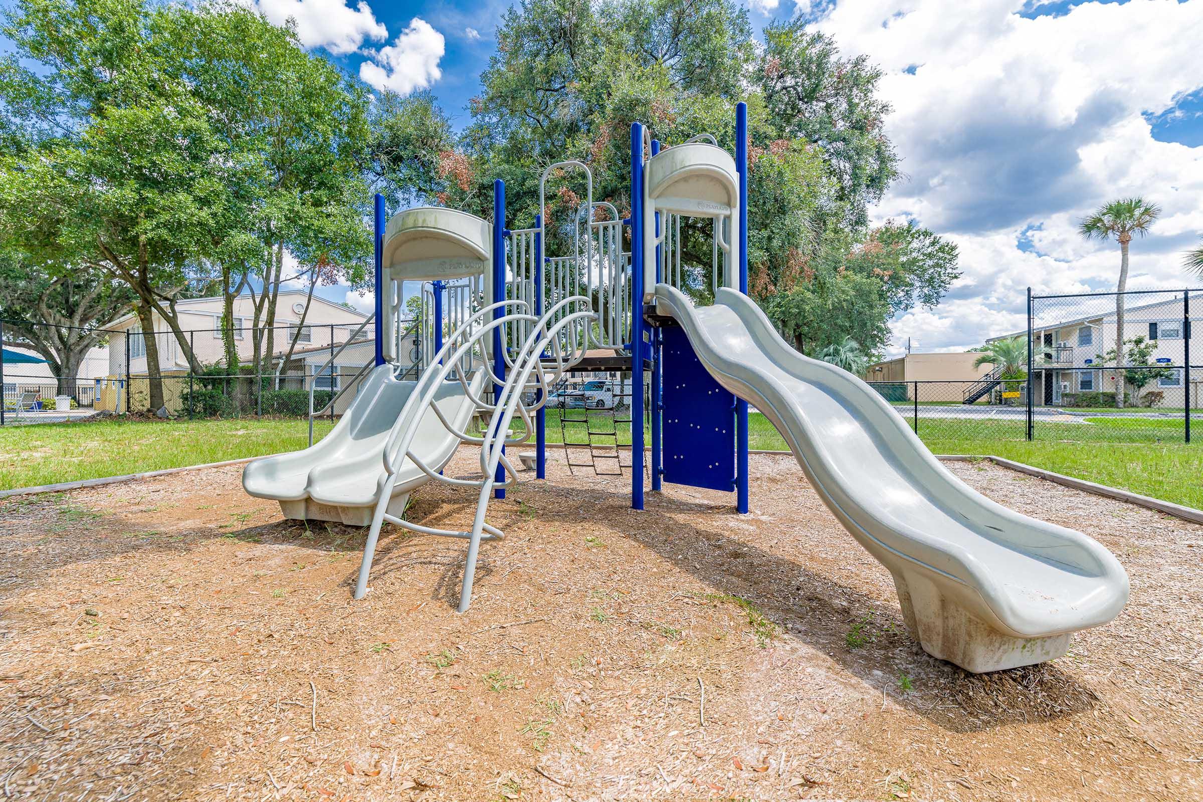 A playground featuring two curved slides, a climbing structure, and surrounded by grass and trees. The sky is partly cloudy, creating a bright atmosphere. In the background, there are residential buildings and a fence, completing the playground scene.