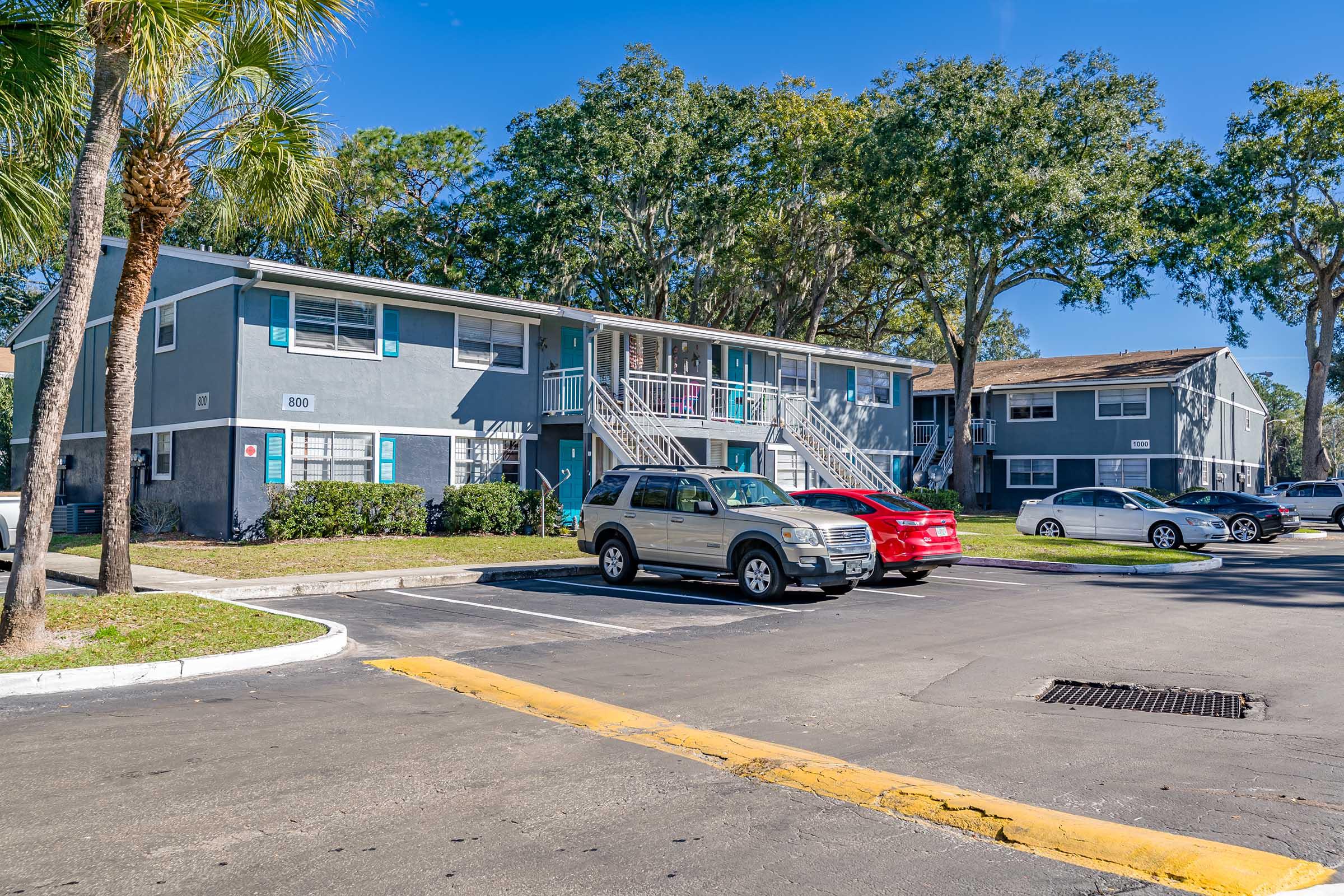 A residential apartment complex featuring two buildings surrounded by palm trees. Parking spaces are visible in front, with several cars parked, including a silver SUV and a red car. The sky is clear and blue, indicating a sunny day.