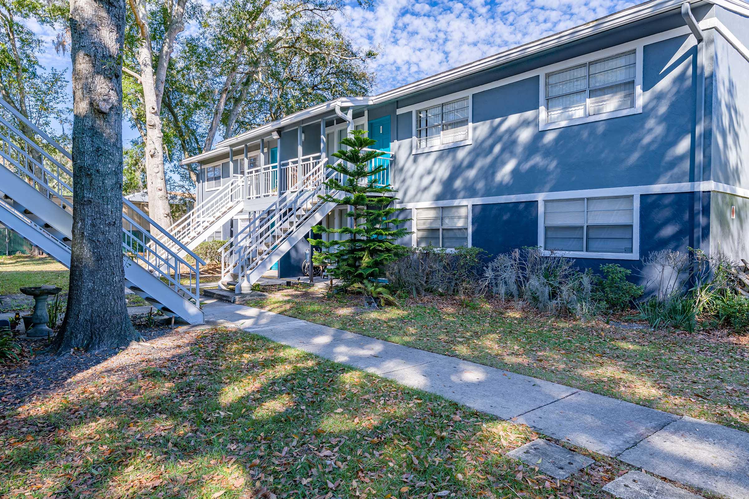 Exterior view of a two-story apartment building with blue siding, surrounded by greenery and leaf-strewn ground. A small staircase leads to the second floor. A unique conifer tree stands in the front yard, and the sky is partly cloudy, creating a bright atmosphere.