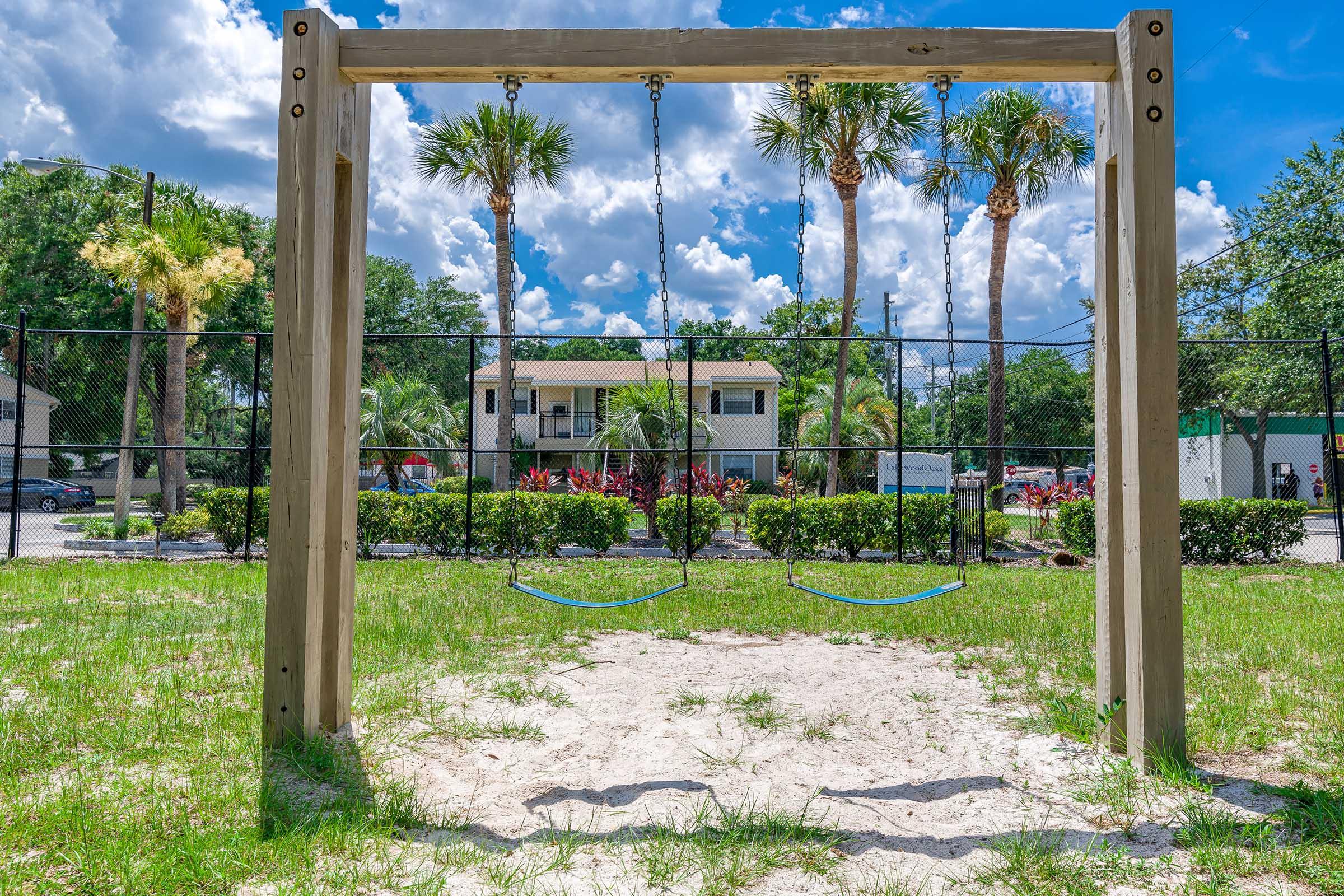 A wooden swing set with two swings hanging from it, set in a grassy area. In the background, there are tall palm trees and a residential building with a white facade. The sky is partly cloudy, creating a sunny atmosphere. The area appears well-maintained, with shrubs and flowers in the foreground.