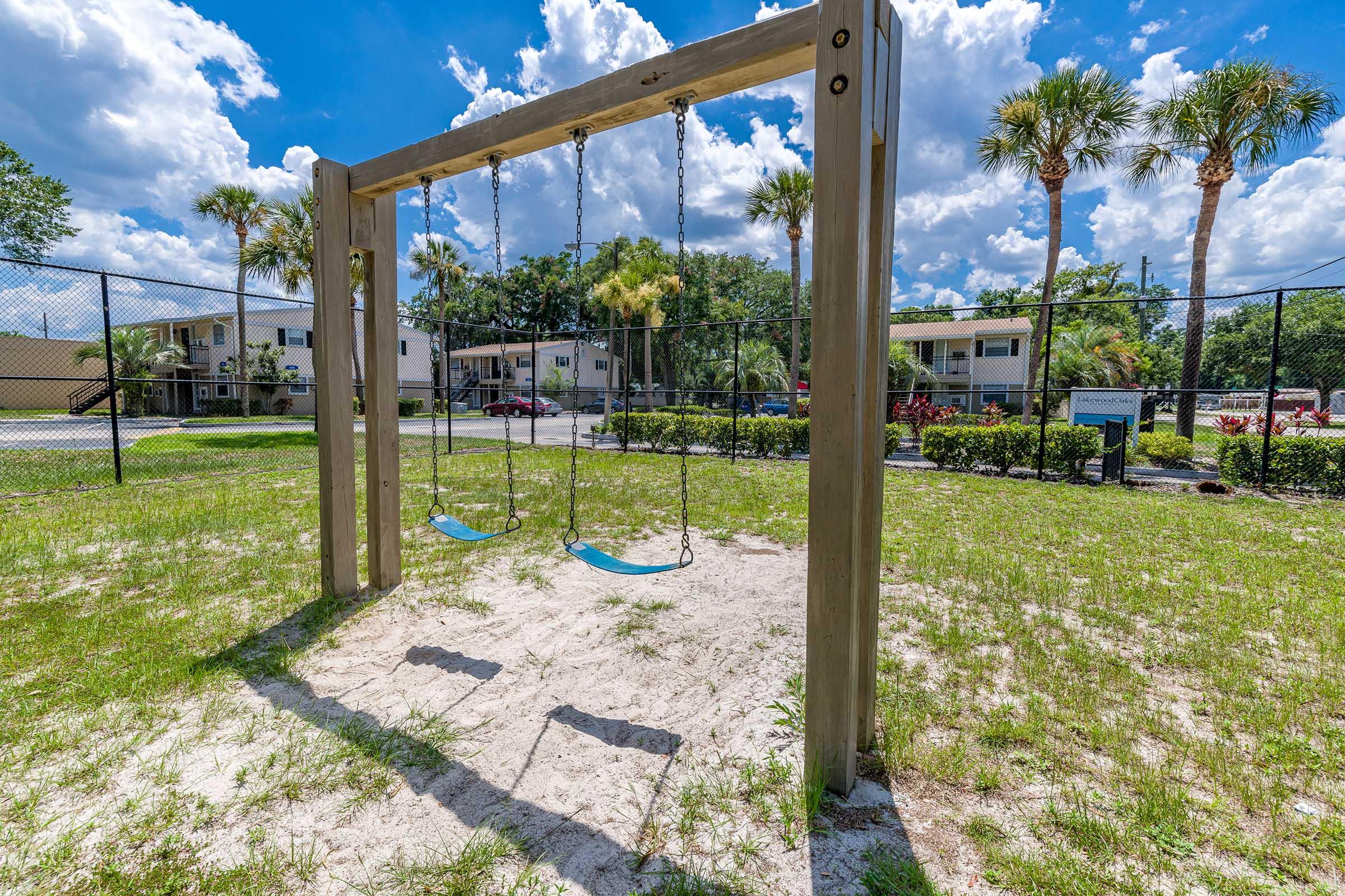 A playground swing set with two swings, situated on a sandy area surrounded by green grass. In the background, there are palm trees and residential buildings under a bright blue sky with fluffy white clouds. A fence encloses the playground area.