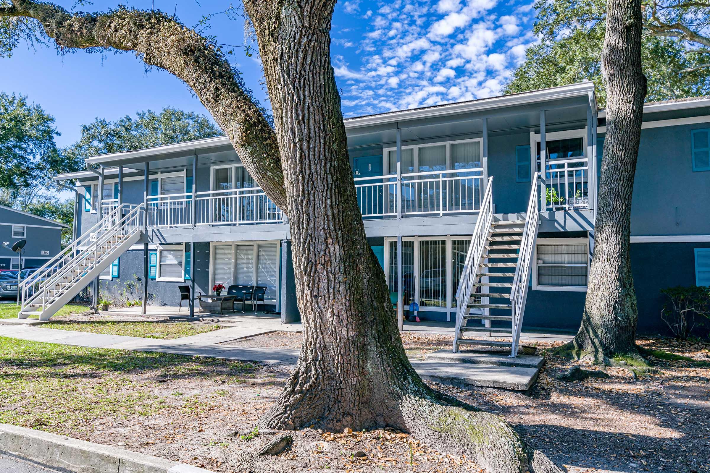A two-story apartment building with a blue facade and white railings. Stairs on either side lead to the second floor, which features balconies. A large tree with a thick trunk is in the foreground, and the area is sunny with a clear blue sky and scattered clouds.
