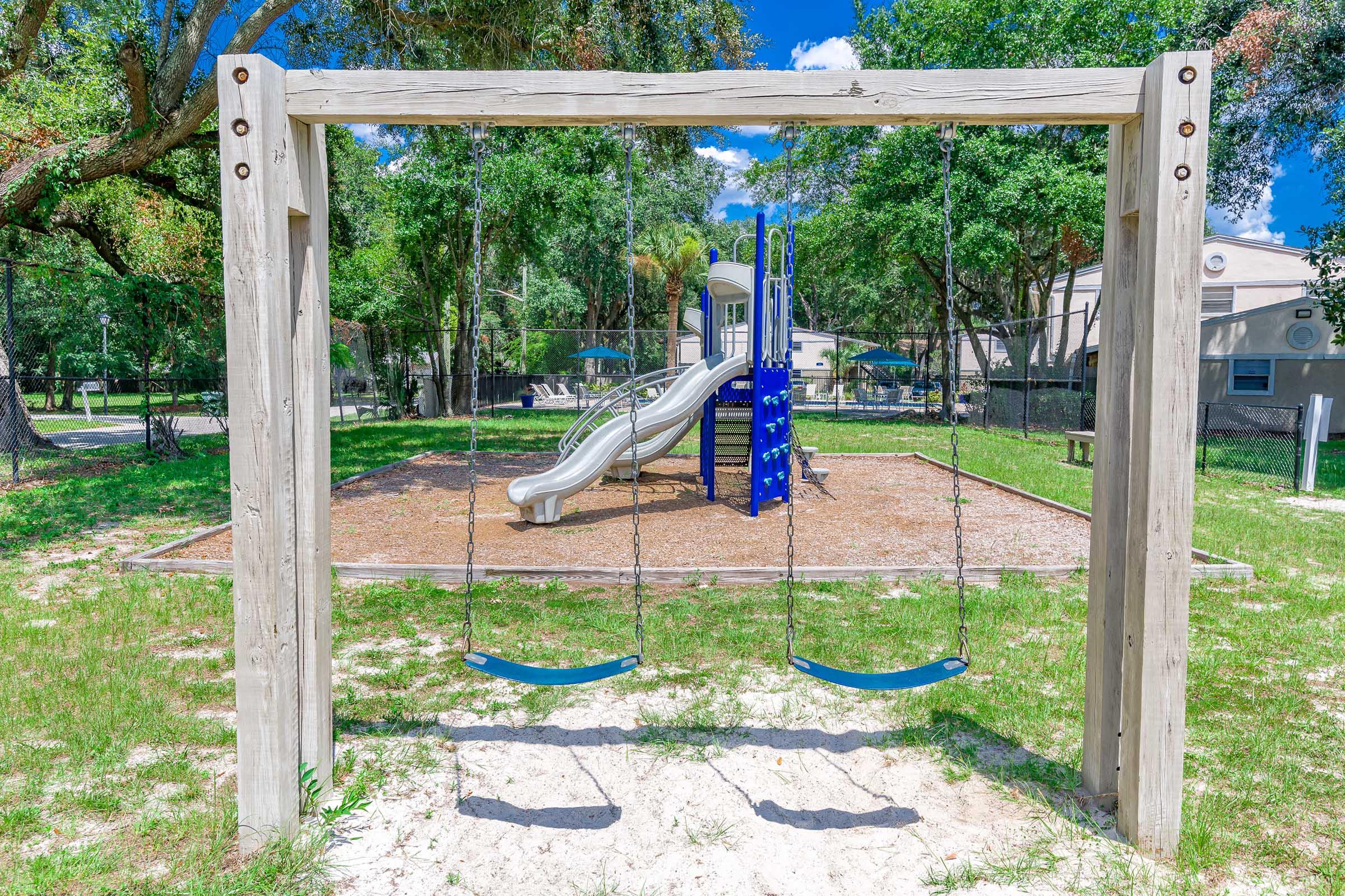 A playground scene featuring wooden swings in the foreground and a blue slide structure in the background, surrounded by grassy areas and trees. The sky is clear with a few clouds, creating a bright and inviting atmosphere.