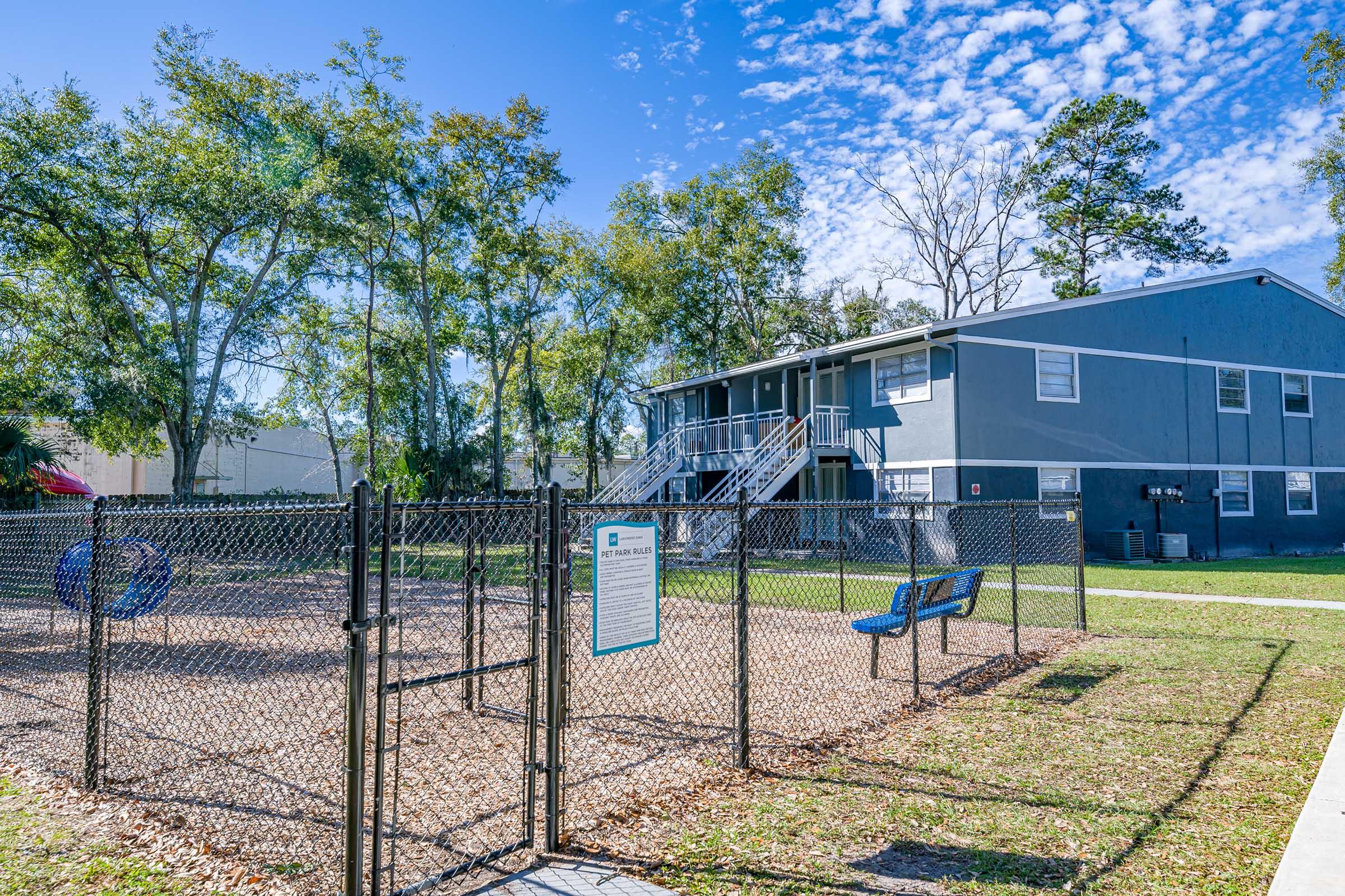 A fenced playground area with two blue swings and a sign, surrounded by trees and grass. In the background, there is a two-story gray building under a partly cloudy sky. The scene is bright and inviting, suitable for children to play.
