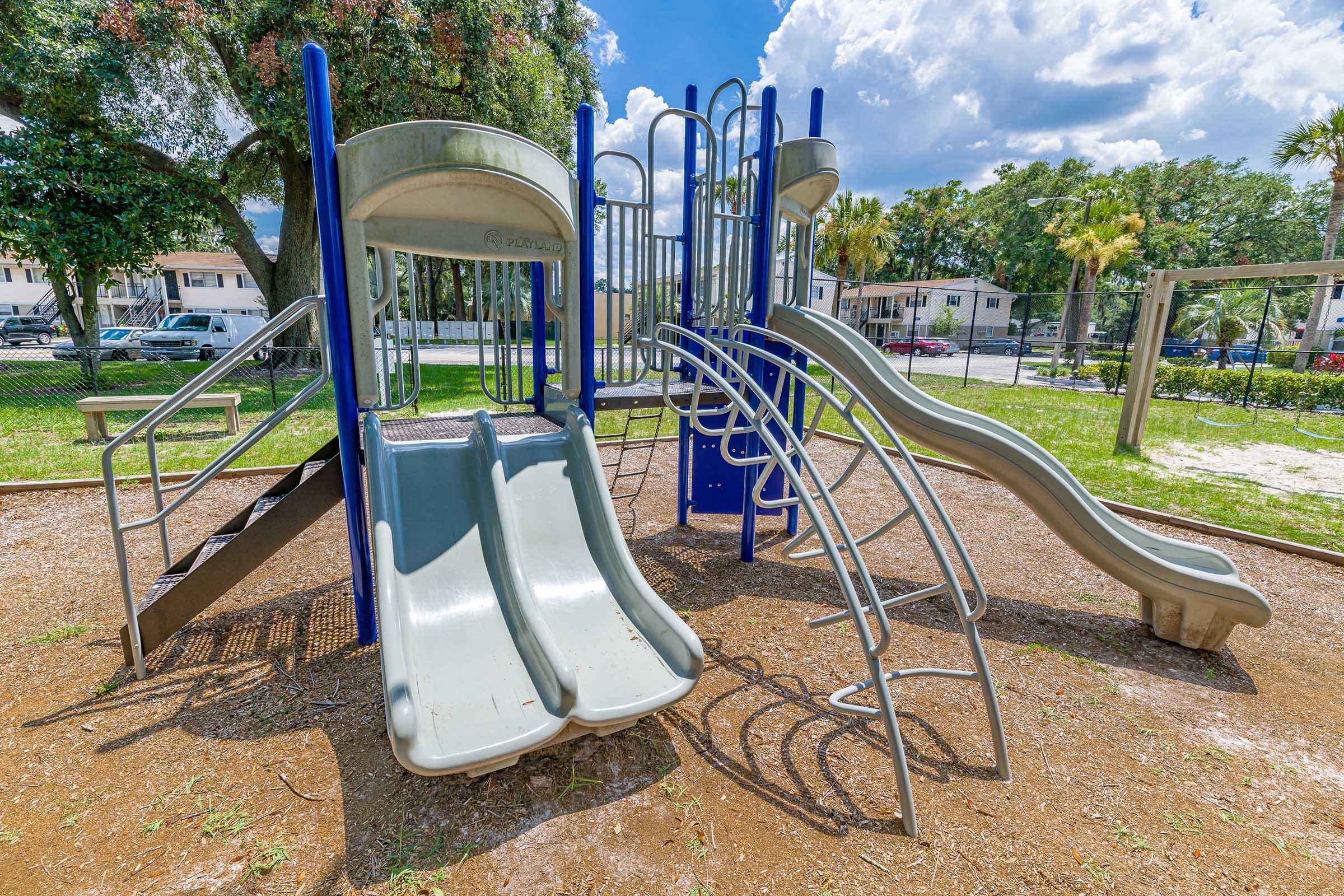 A colorful playground featuring two slides and climbing structures, set in a grassy area with trees in the background. The sky is partly cloudy, and there are nearby residential buildings.