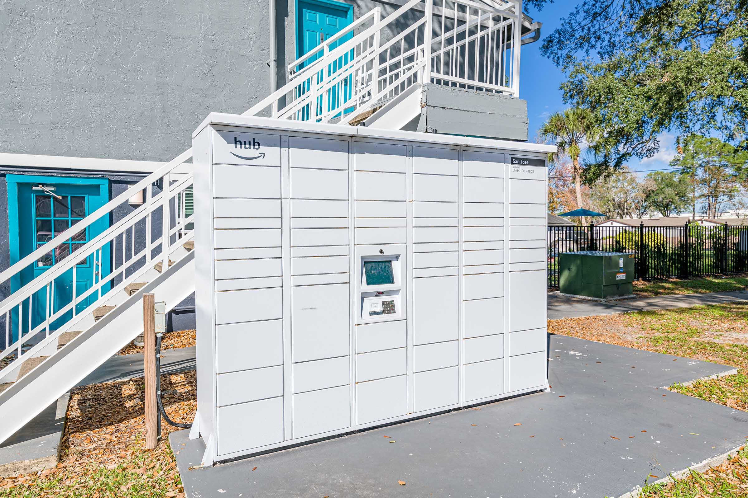 A white Amazon package locker situated outdoors near a building. The locker features multiple compartments for package deliveries and has a digital interface for retrieving items. Surrounding the locker are landscaped areas with fallen leaves and a staircase leading to an upper level of the building.