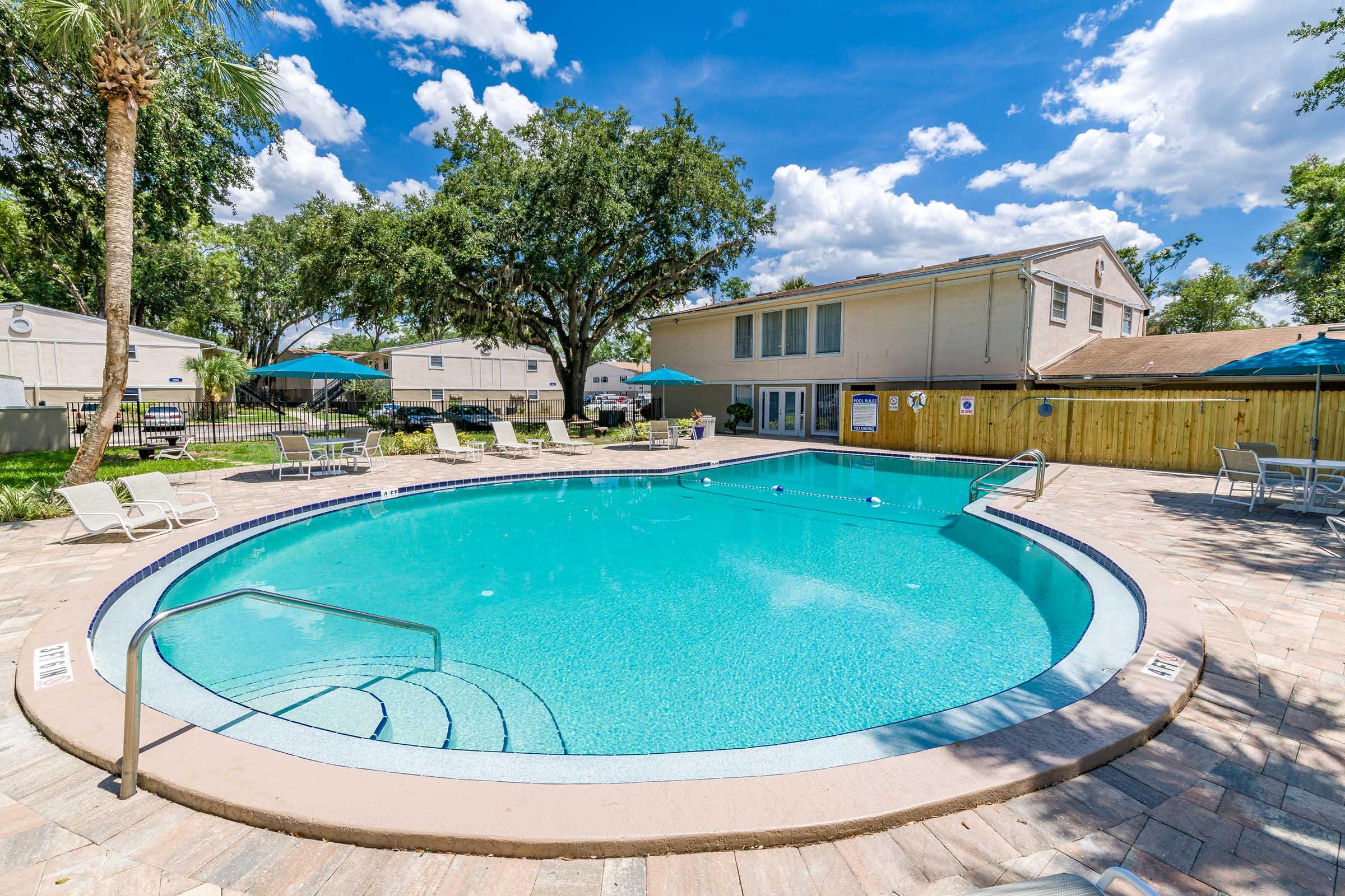A sparkling blue swimming pool surrounded by lounge chairs, with vibrant green grass and palm trees. In the background, a light-colored building and a wooden fence can be seen under a bright blue sky with fluffy white clouds. Umbrellas provide shade for relaxation.