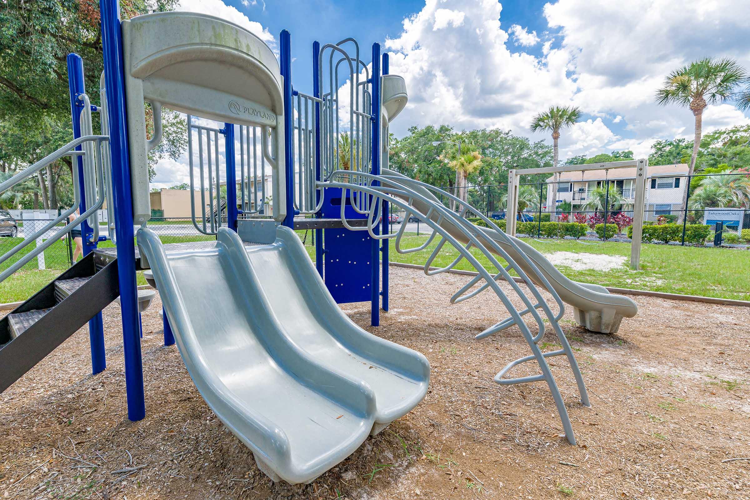 A playground featuring two gray slides and climbing structures set on grass. The background includes palm trees and a residential area under a bright, partly cloudy sky.