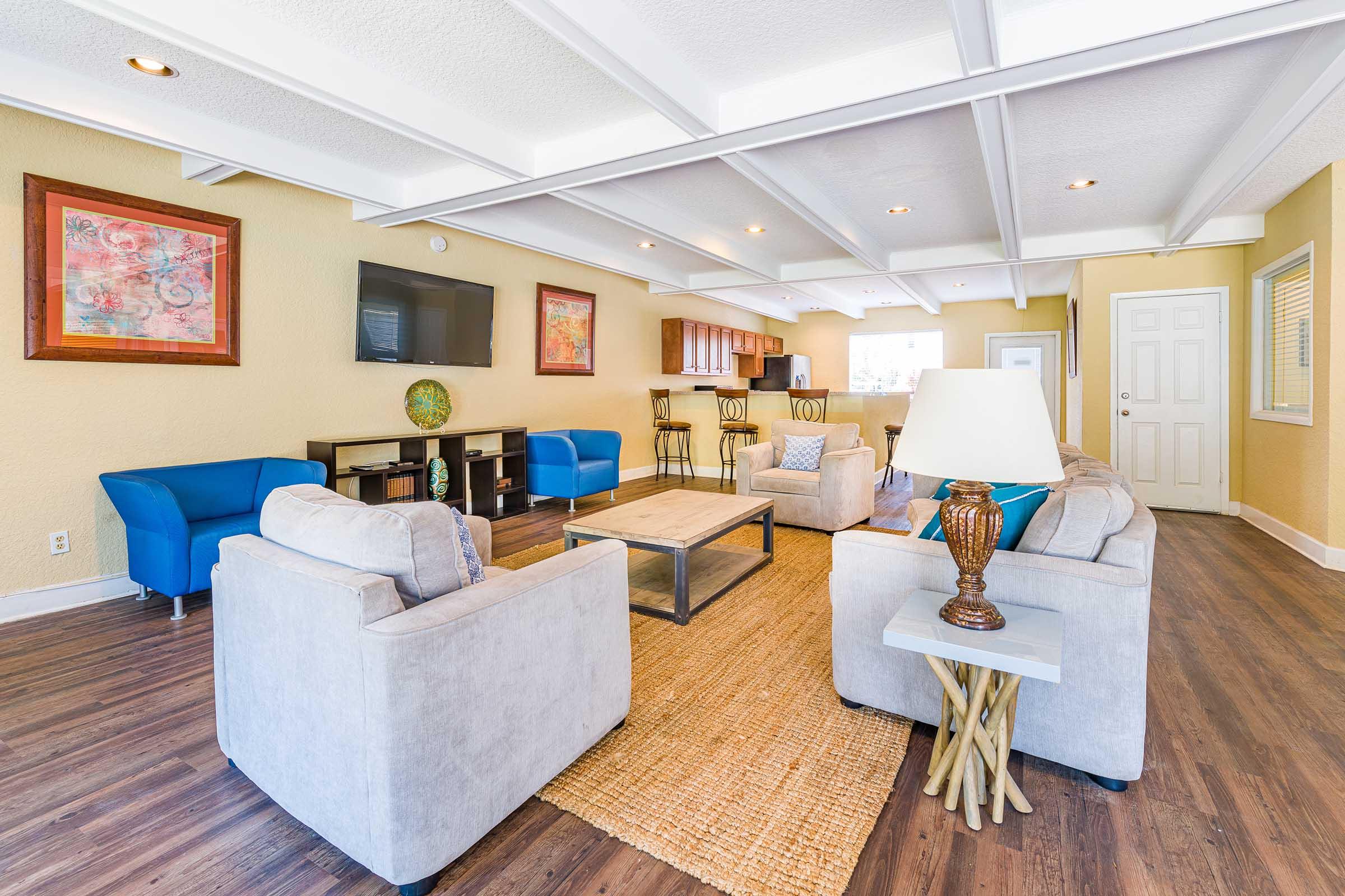 Bright and spacious living area featuring gray sofas and blue chairs, a wooden coffee table, and a decorative lamp. The room has a light color scheme with exposed beams and artwork on the walls. A bar area with stools is visible in the background, along with a TV and doors leading to another space.