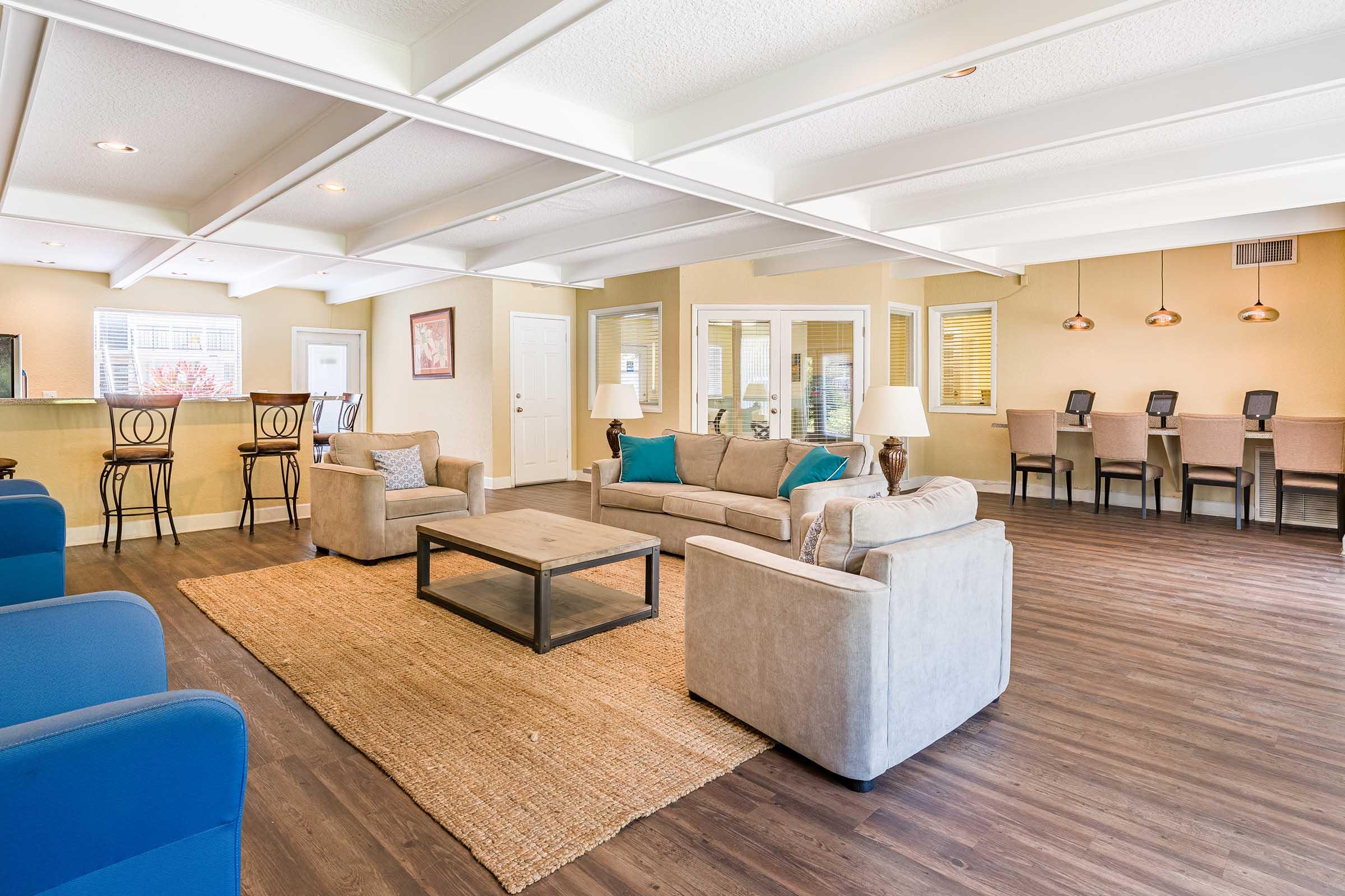 A spacious, modern living area featuring a light-colored sofa with blue cushions, an area rug, and a coffee table. Chairs are arranged around a dining area in the background, complemented by large windows that allow natural light. The walls are painted in warm tones, creating an inviting atmosphere.