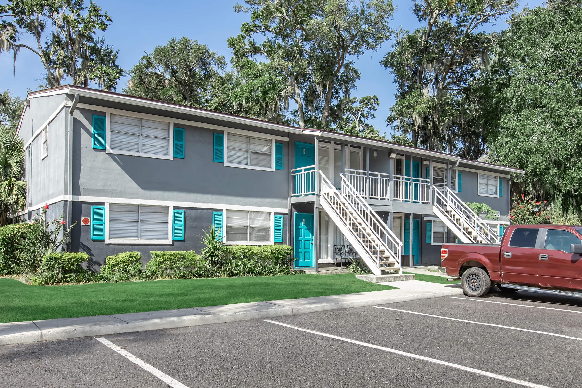 A two-story apartment building with gray exterior and teal accents, featuring multiple windows and stairs leading to the second floor. In front, there is a red pickup truck parked in a lot, with green landscaping and trees in the background.